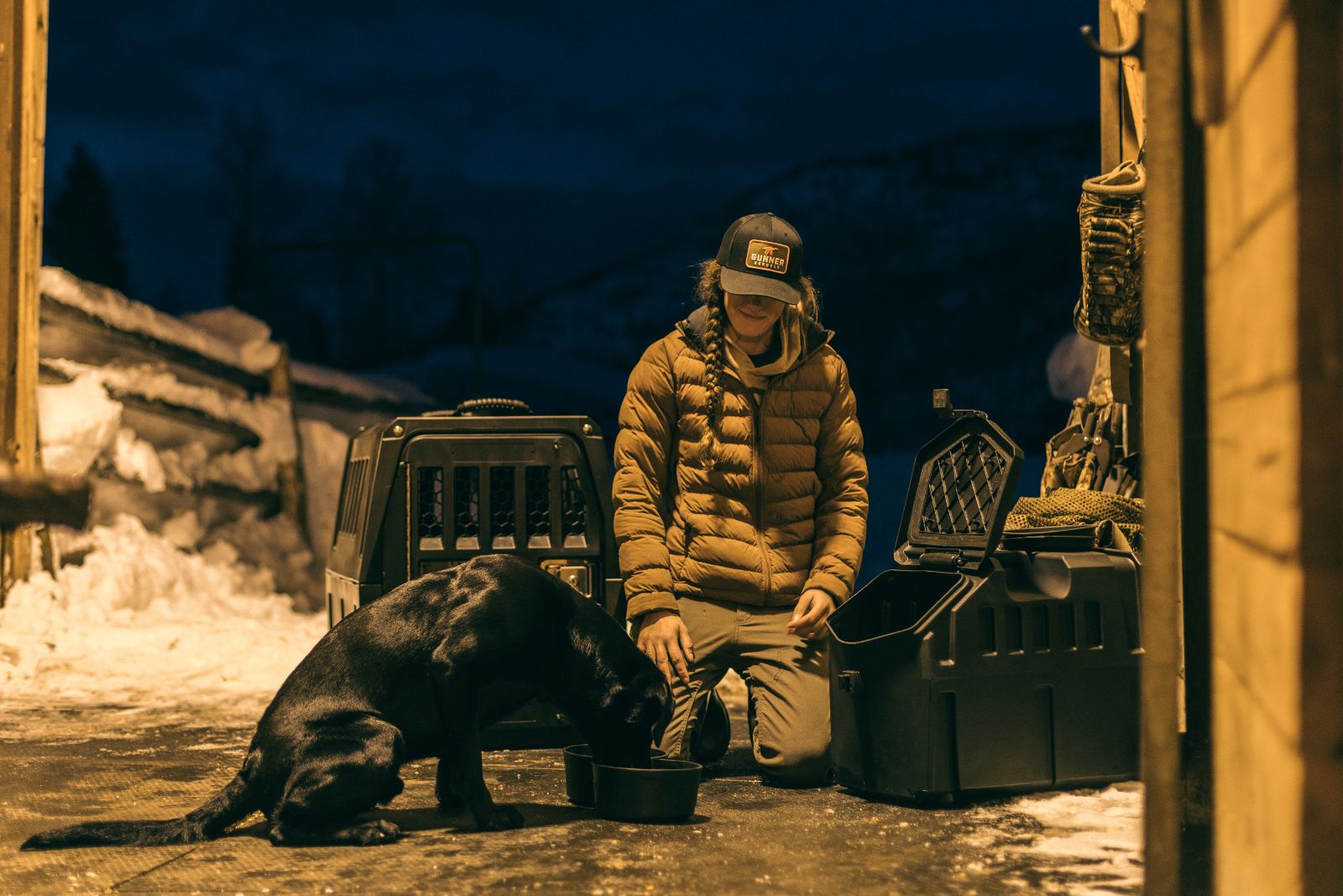 A person in a brown jacket and cap kneels beside a black dog eating from a bowl near pet carriers, outdoors at night with snow and mountains in the background.