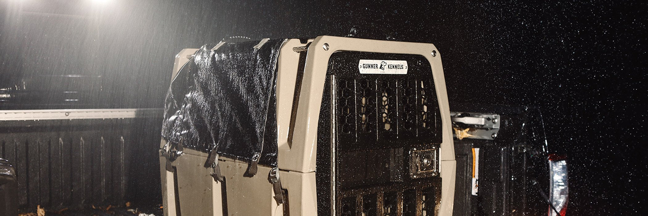A tan animal crate sits in the bed of a pickup truck at night in the rain, illuminated by the truck’s lights. The crate is partially covered with a wet black tarp.