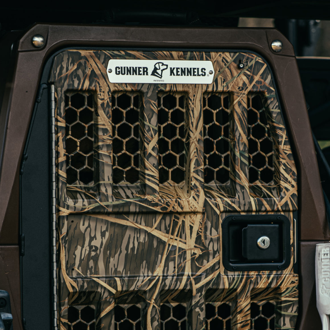 Close-up of the G1™ KENNEL in Susquehanna/Mossy Oak Shadow Grass by GUNNER, featuring hexagonal vents, a black latch, and a metallic plate with the GUNNER dog logo—perfect for duck and goose hunters.