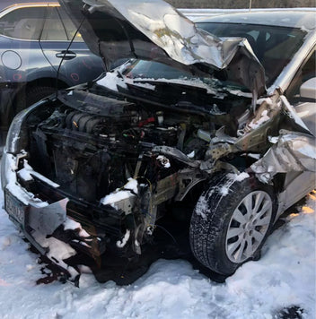 A silver car with severe front-end damage and an exposed engine is parked on snow, showing signs of a recent accident. Nearby, a working dog sniffs around the wreckage while a dog crate sits beside the crumpled hood.