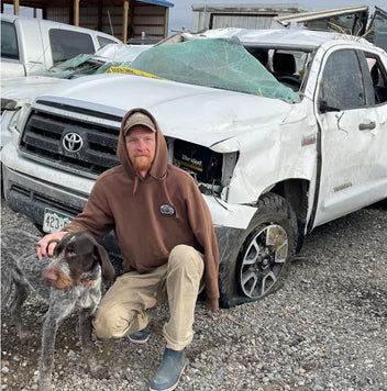 A man in a brown hoodie kneels next to his gray hunting dog in front of a badly damaged white Toyota pickup truck with a smashed windshield and dented body, parked among other vehicles.