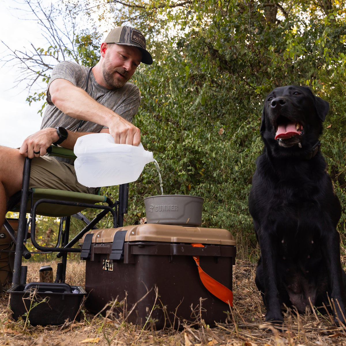 A man wearing a BOTTOMLAND MALLARD TRUCKER hat sits in a folding chair, pouring water from a jug into a bowl on top of a cooler next to a black Labrador.