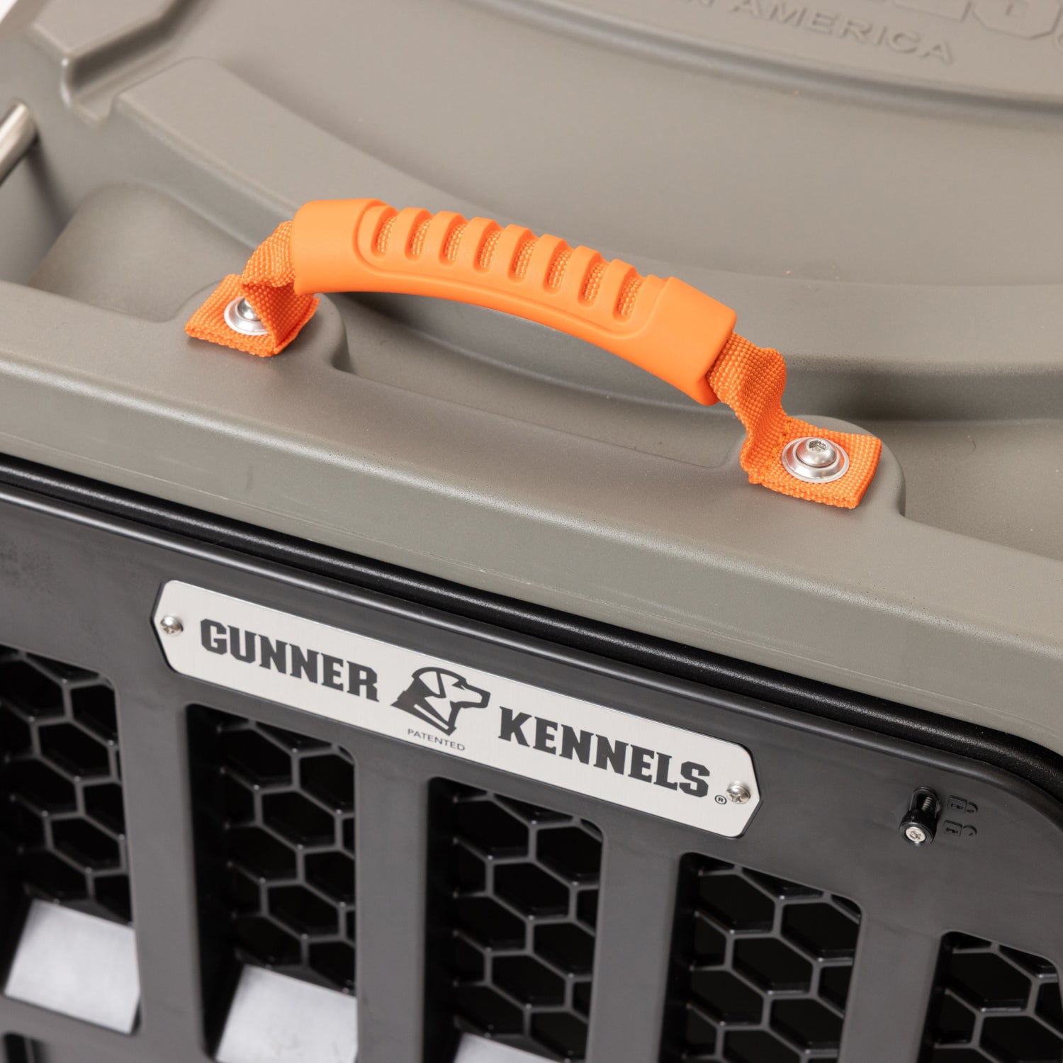 Close-up of a GUNNER dog crate equipped with KENNEL HANDLES - BLAZE ORANGE, featuring a ridged rubber grip and a metal label with the GUNNER brand name and logo, attached to a robust gray and black crate built from GUNNER-Grade materials and highlighted by its signature hexagonal vent pattern.