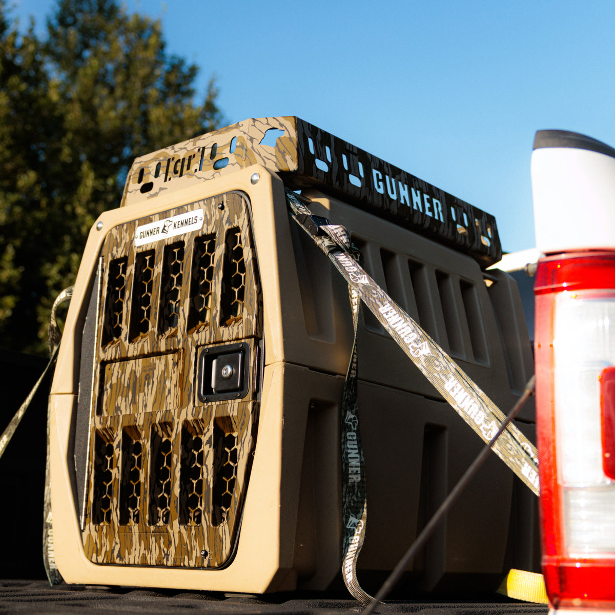 A GUNNER Kennel Rack in Mossy Oak Bottomland, featuring ventilation holes and the GUNNER logo, is secured in the bed of a truck with sturdy latches and straps—maximizing your truck bed space. With trees and blue sky in the background, this kennel storage system keeps your gear organized for every adventure.