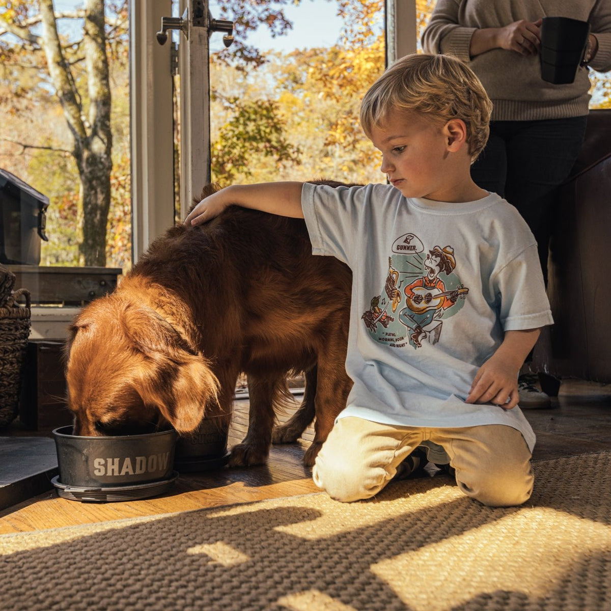 A young boy in a DOG & DUCK BAND TEE by GUNNER gently pats his golden retriever, Shadow, eating from a bowl as sunlight streams in. An adult with a mug stands behind him, and a Dog & Duck Band poster is on the wall.