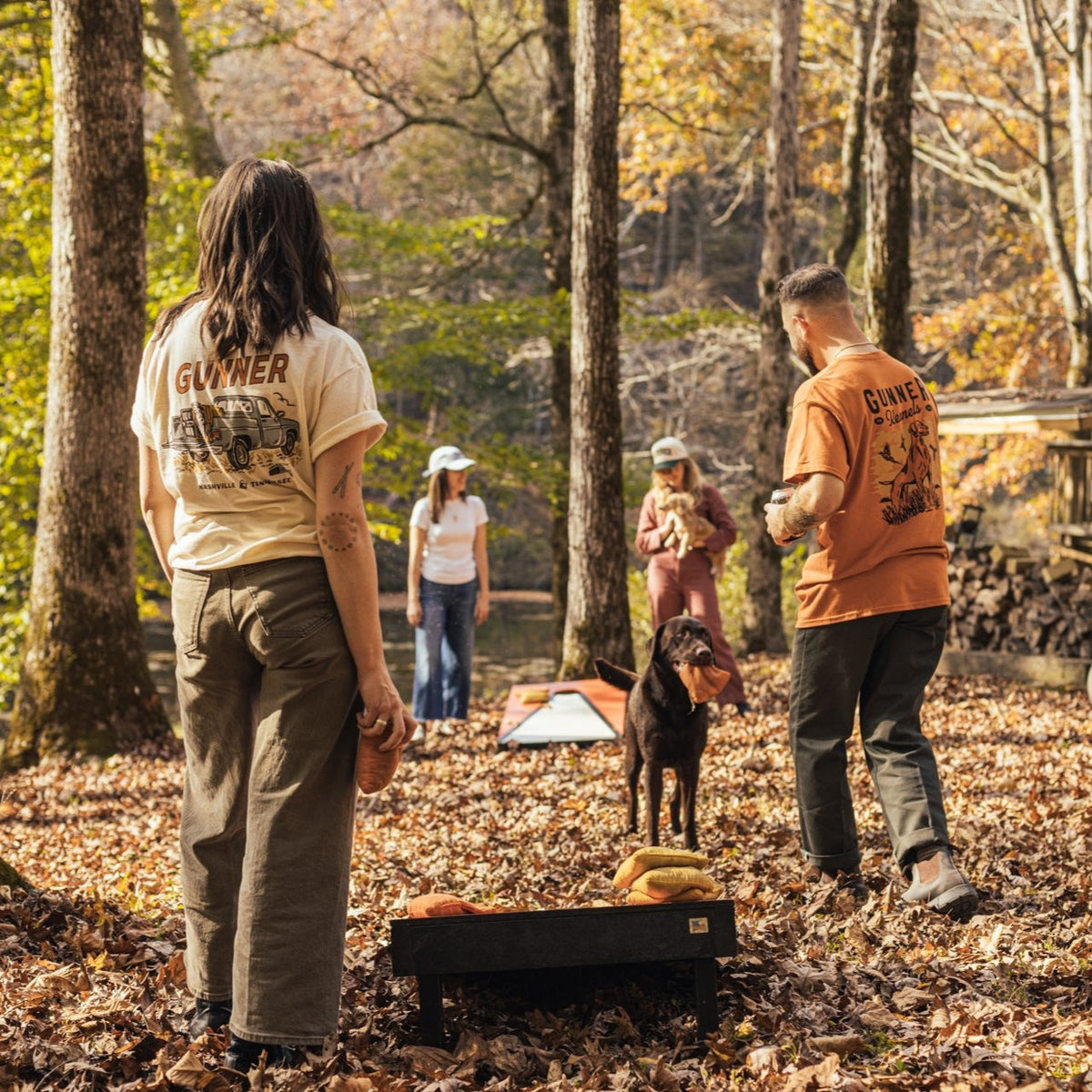 A group enjoys outdoor cornhole among autumn leaves, a dog on point near the boards. Some wear GUNNER UPLAND TEEs as sunlight filters through the forest.