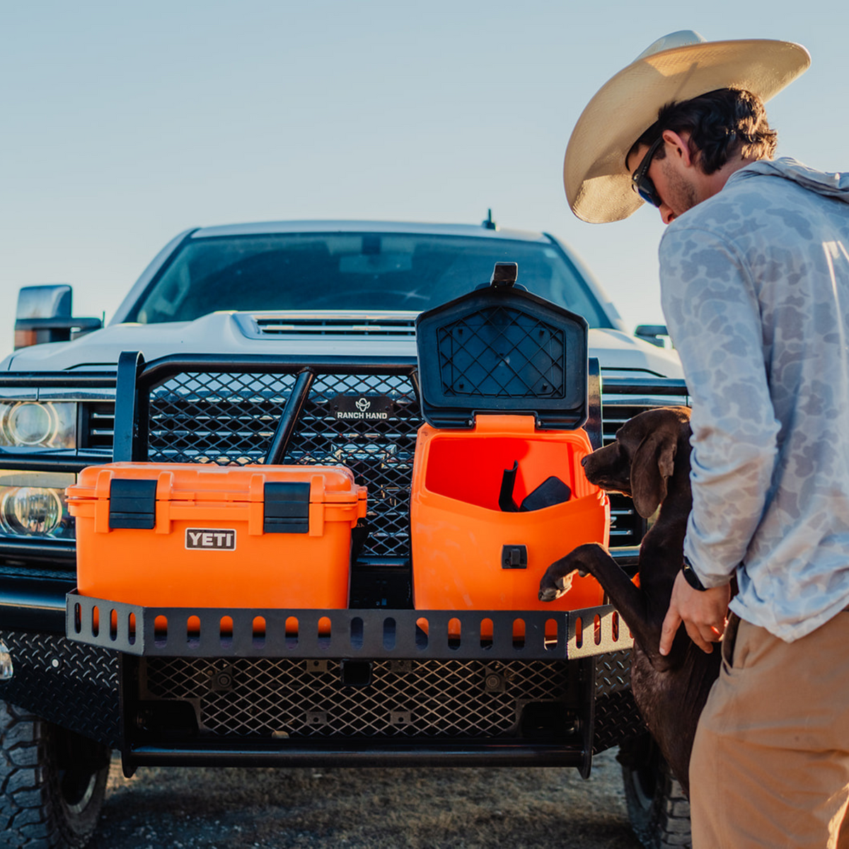 A man in a cowboy hat stands by a parked truck, holding a brown dog reaching into an open, limited-edition GUNNER FOOD CRATE - BLAZE ORANGE by GUNNER—perfect for dog food storage. Another closed GUNNER crate sits beside it.