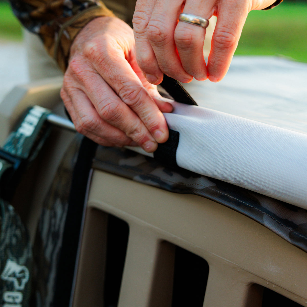 Wearing a gold ring, a person adjusts the black strap on the GUNNER ALL-WEATHER KIT 2.0 MOSSY OAK BOTTOMLAND, a tan pet crate outdoors that’s covered with UV-protective white fabric, with green grass in the background.