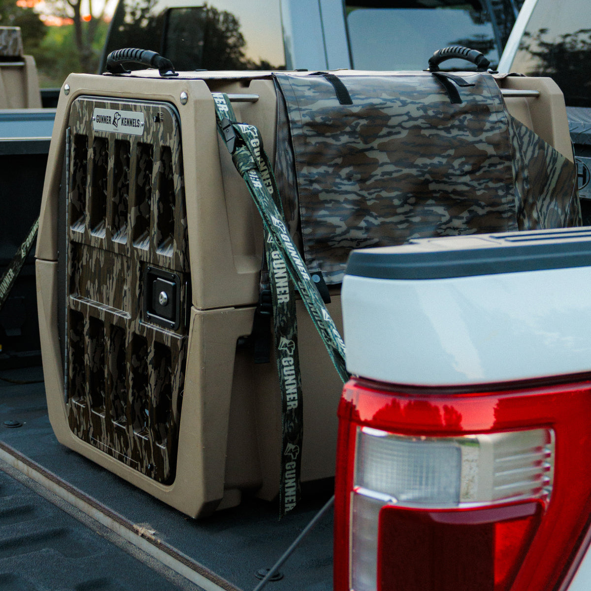 A GUNNER ALL-WEATHER KIT 2.0 MOSSY OAK BOTTOMLAND dog kennel, featuring a durable camouflage cover that offers UV protection and waterproofing, is secured with straps in the bed of a pickup truck. The tailgate is open, and the kennel includes ventilation openings for added comfort.