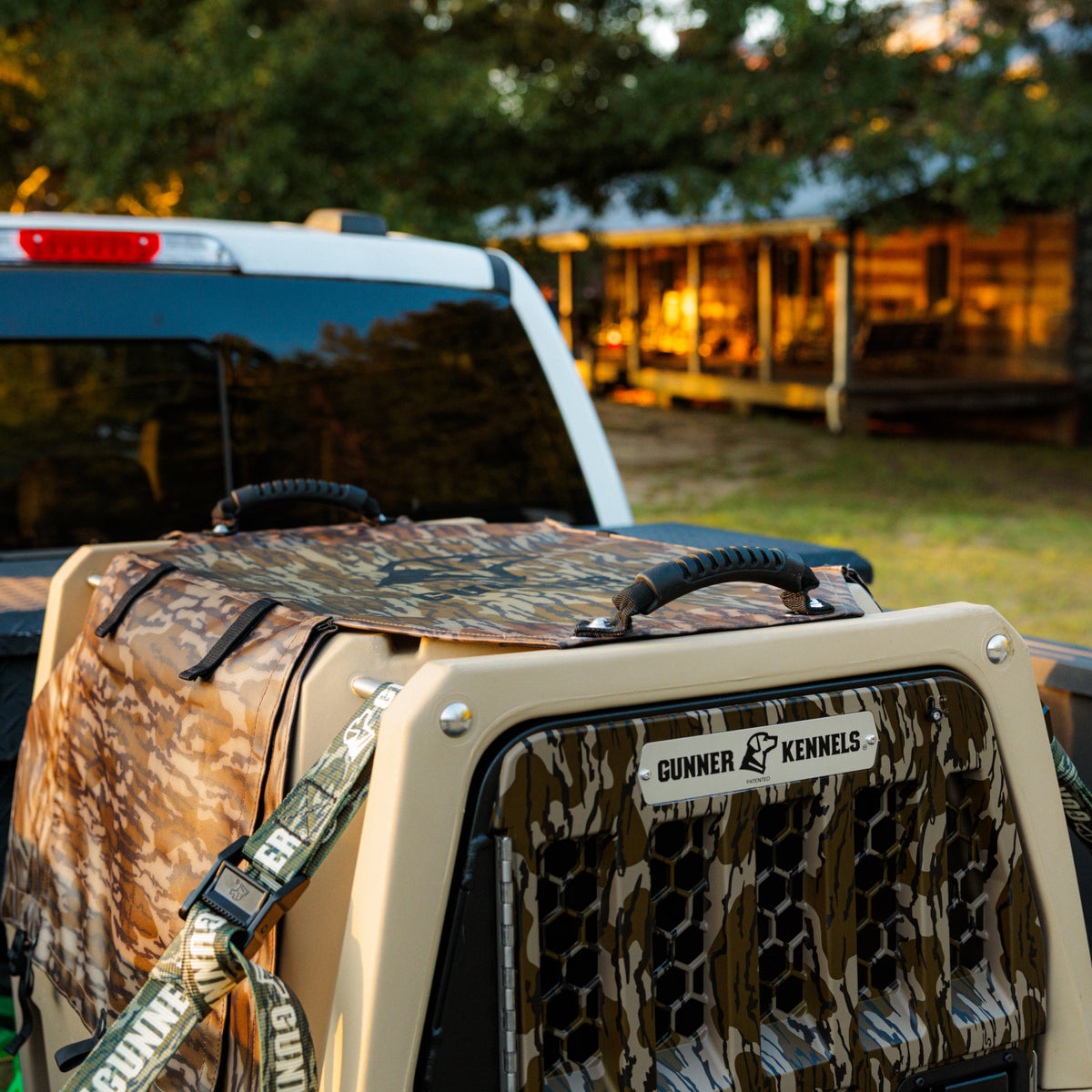 A GUNNER kennel outfitted with the ALL-WEATHER KIT 2.0 MOSSY OAK BOTTOMLAND is safely secured in the bed of a pickup truck, parked outside a cabin surrounded by trees and warm sunlight, providing superior UV protection and enhanced durability for your dog.