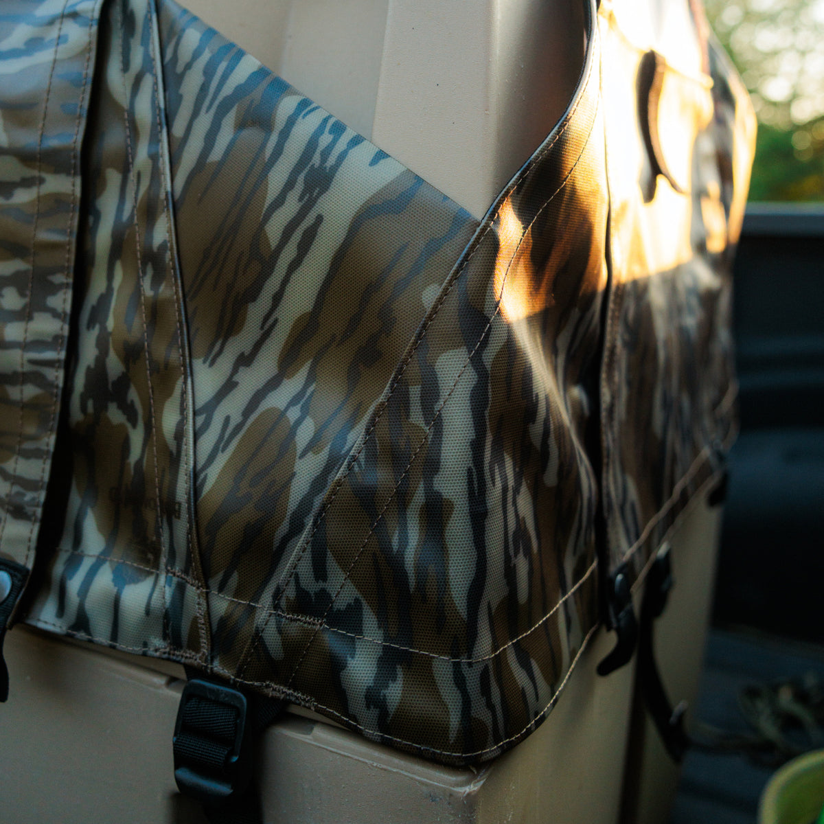 A close-up of the GUNNER ALL-WEATHER KIT 2.0 MOSSY OAK BOTTOMLAND shows its camouflage fabric draped over a vehicle’s back seat, featuring sturdy black straps and buckles. Sunlight highlights the distinctive pattern and rugged construction of this waterproof cover, which provides dependable UV protection—making it an excellent all-weather option for your dog kennel.