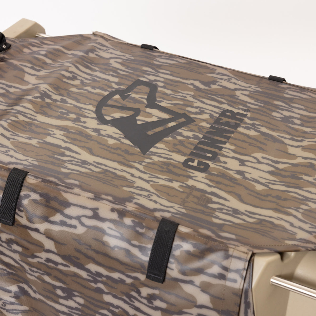 A close-up of a tan and brown Mossy Oak camo-patterned fabric cover with a prominent black GUNNER logo and text, secured to a hard surface with black straps. The ALL-WEATHER KIT 2.0 MOSSY OAK BOTTOMLAND from GUNNER is a waterproof dog kennel cover, designed for durability, UV protection, and reliable performance during your outdoor adventures.