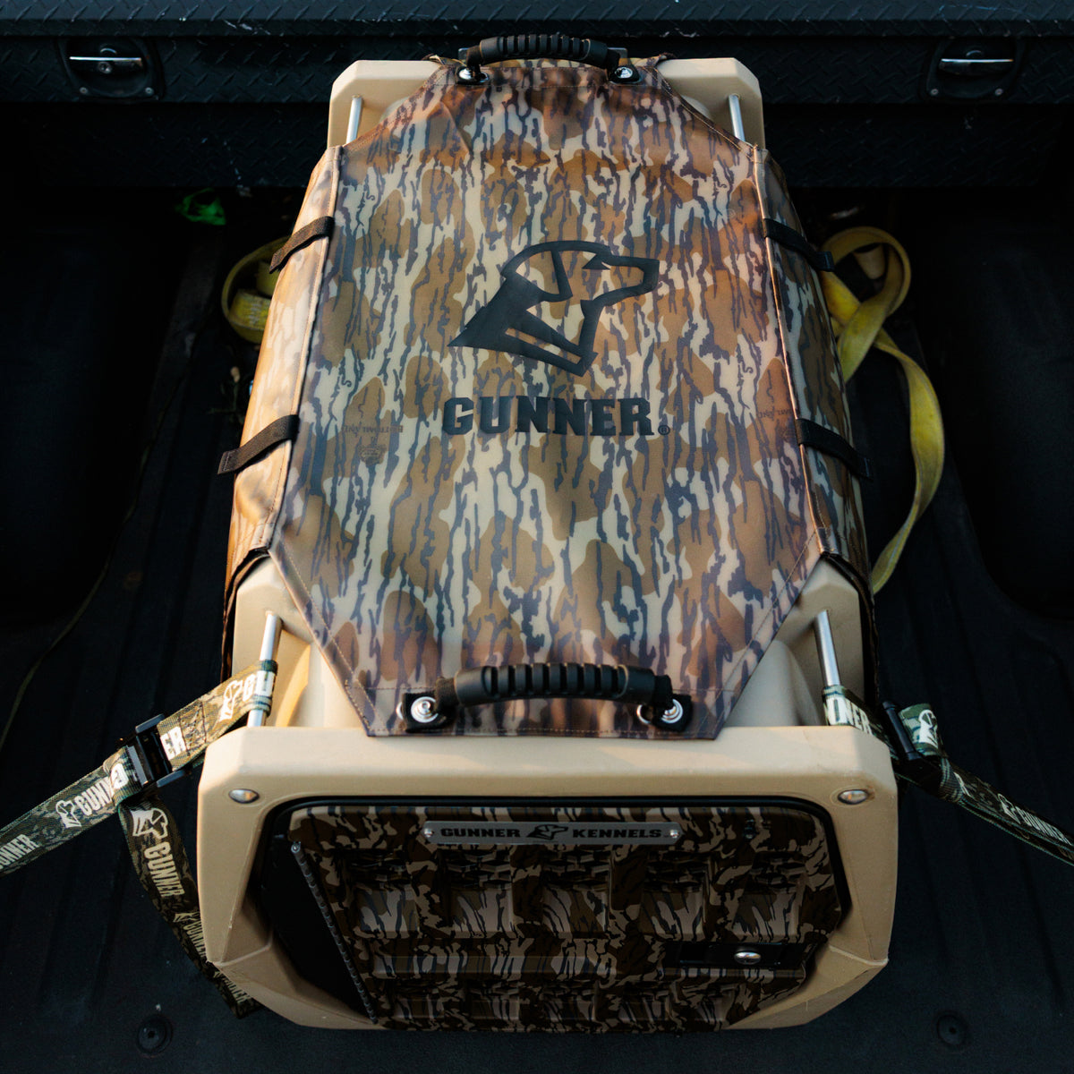 A tan dog crate labeled GUNNER is situated in the back of a vehicle, fitted with the ALL-WEATHER KIT 2.0 MOSSY OAK BOTTOMLAND—a camouflage-patterned, waterproof kennel cover and straps from GUNNER. The cover provides dependable UV protection, making it perfect for outdoor adventures.