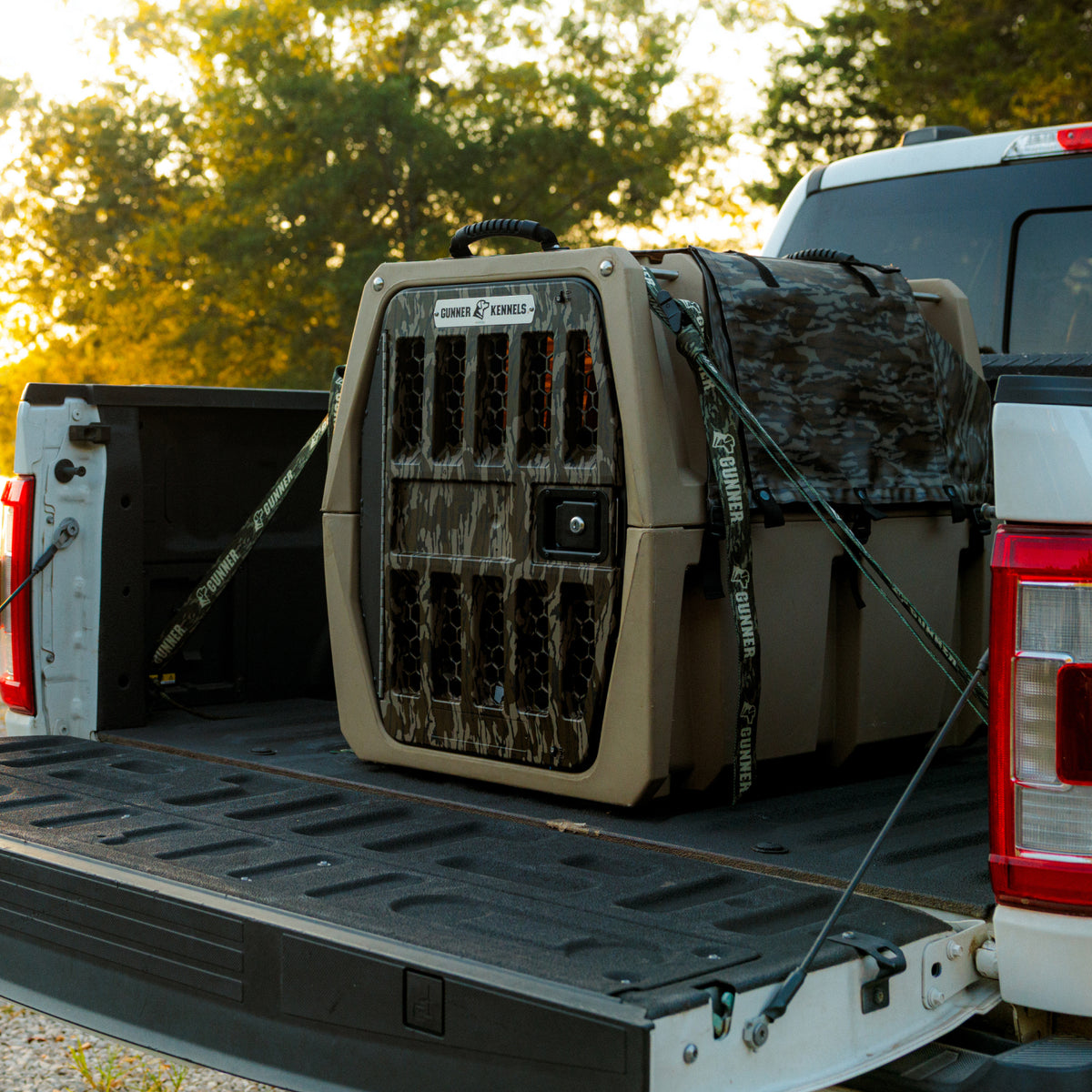 A GUNNER ALL-WEATHER KIT 2.0 MOSSY OAK BOTTOMLAND dog crate, secured with straps and featuring its waterproof cover, sits in the bed of a white pickup truck parked outdoors near trees at sunset.