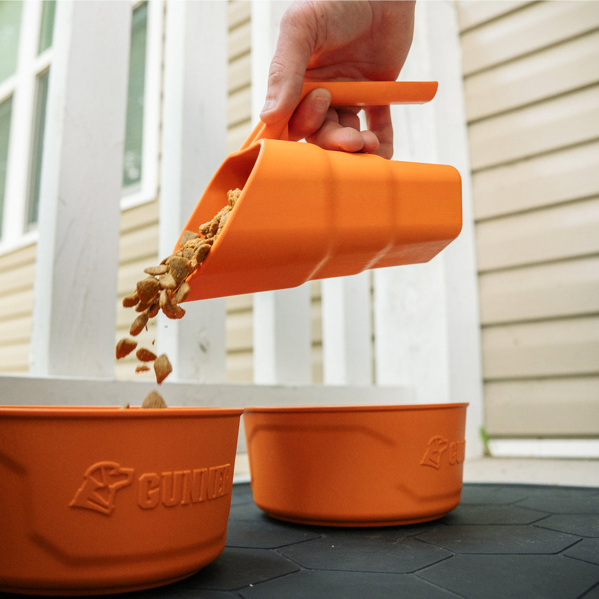 A hand uses an ergonomic FOOD SCOOP BLAZE ORANGE to pour dry pet food into an orange bowl.