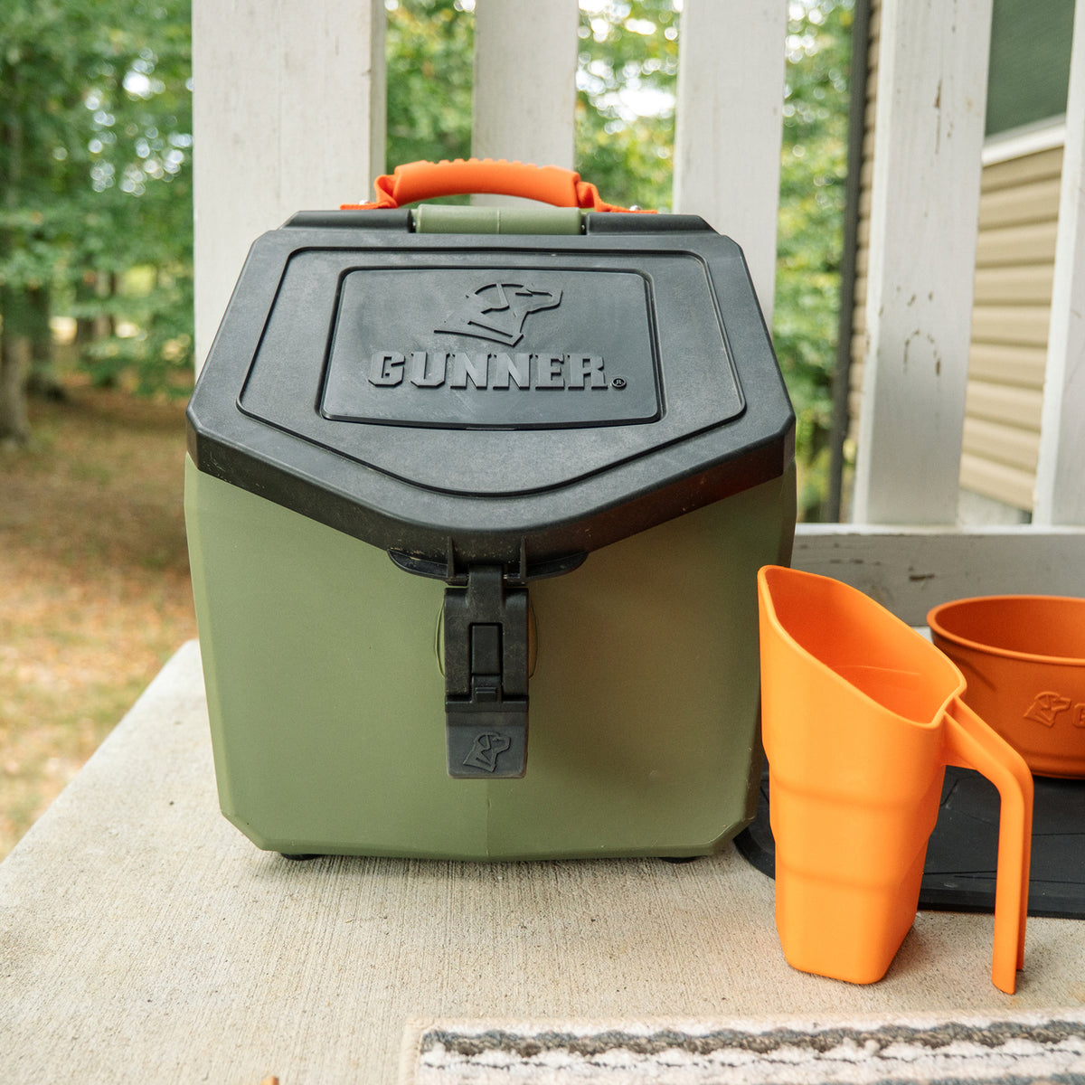 An olive green storage container with a black lid and orange handle, sitting next to a GUNNER FOOD SCOOP BLAZE ORANGE and a small orange bowl.