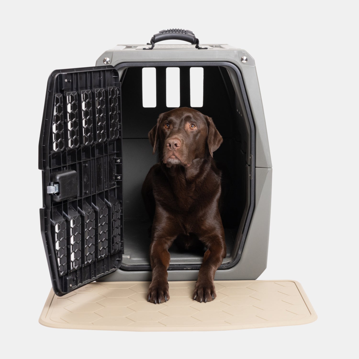 A chocolate Labrador dog sits inside a gray pet crate with the door open, resting its front paws on a GUNNER BOWL MAT - TAN placed outside the crate.