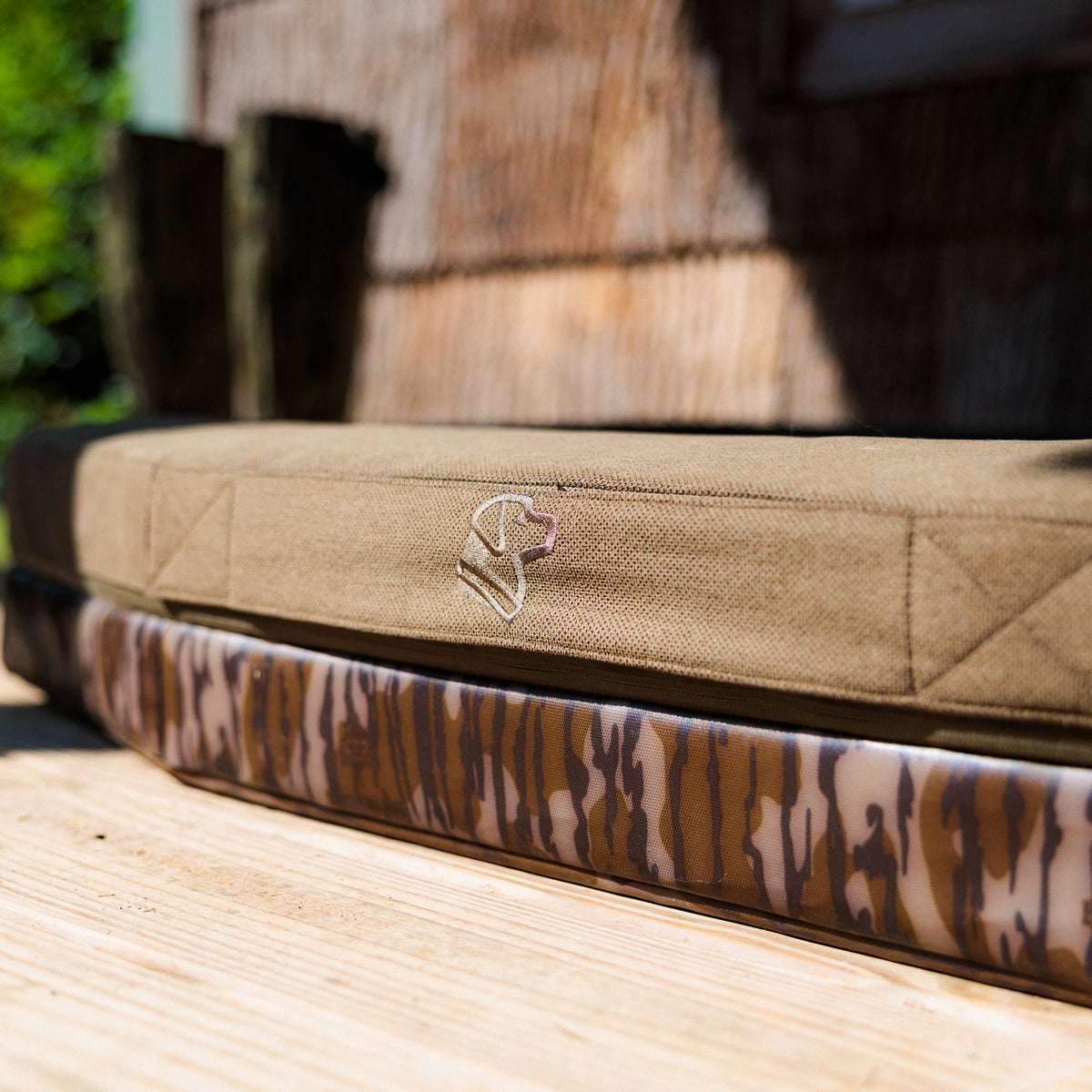 Two GUNNER CAMP BEDs are displayed on a wooden surface outdoors. One is a durable CAMP BED in solid tan with a stitched dog head logo, while the other showcases a brown and tan Mossy Oak Bottomland camouflage pattern. Sunlight and a blurred wooden wall in the background enhance the cozy GUNNER CAMP BED vibe.