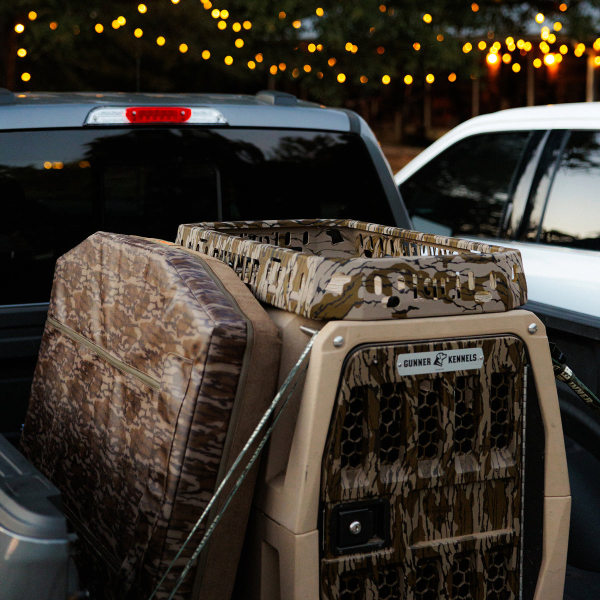 A GUNNER CAMP BED and a camouflage dog crate are secured in the back of a pickup truck at dusk, with string lights glowing in the background.