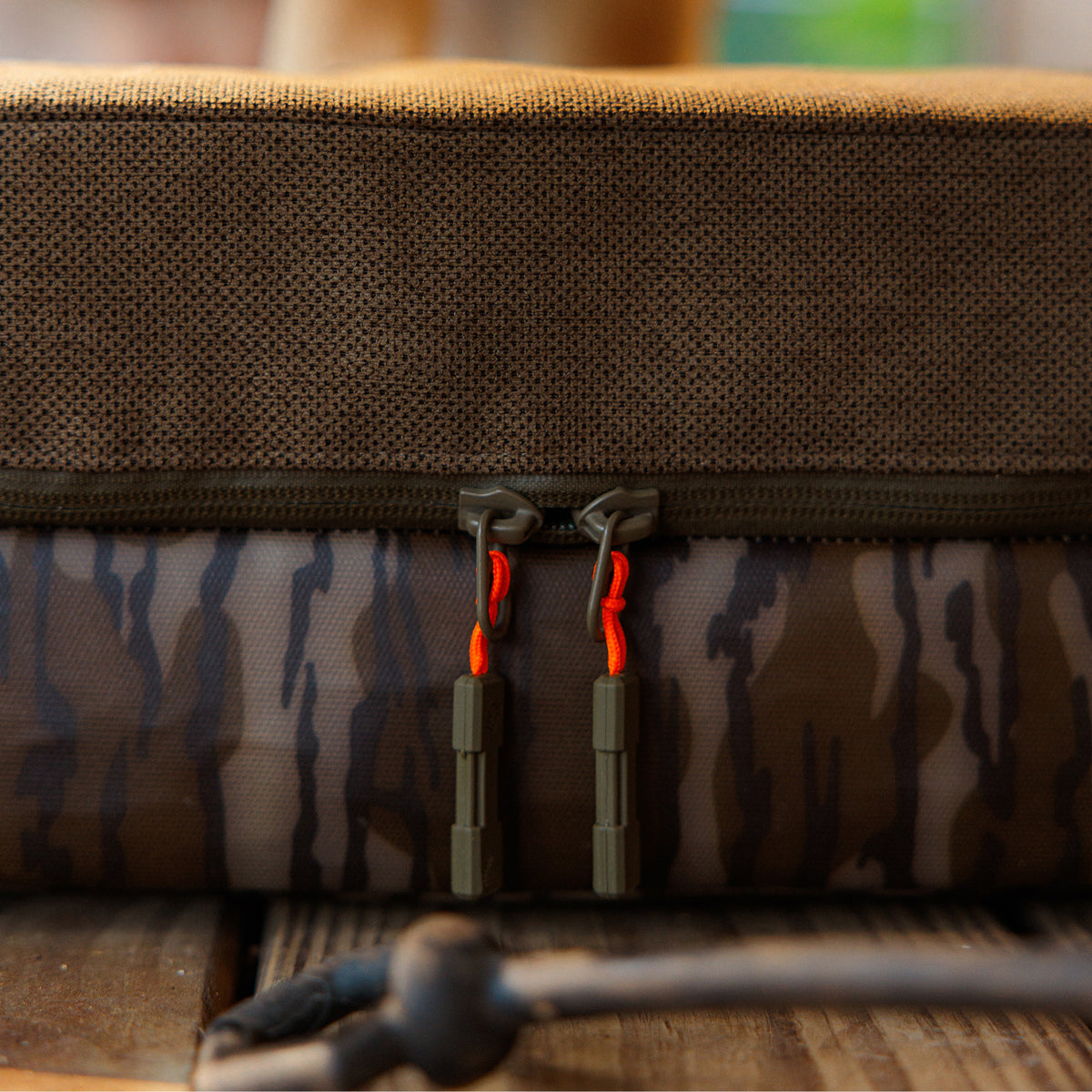 Close-up of GUNNER’s CAMP BED in Mossy Oak Bottomland camo, featuring two brown zipper pulls with bright orange cords, resting on a wooden surface—designed as a tough, adventure-ready dog bed for your companion.