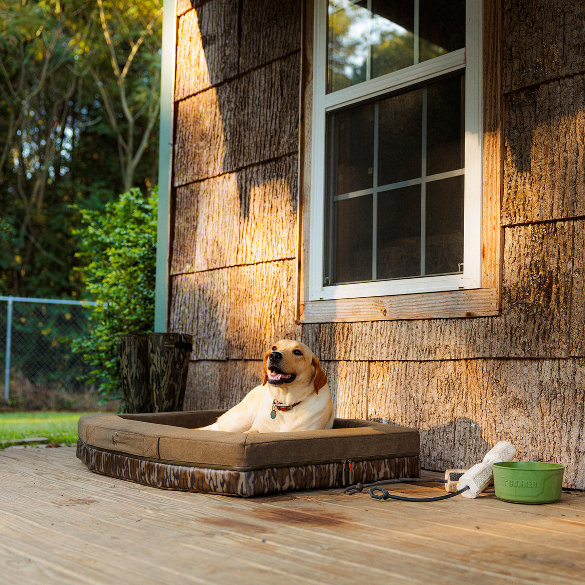 A yellow Labrador retriever relaxes on the GUNNER CAMP BED, a durable dog bed, on a wooden porch next to a green water bowl, a leash, and a toy, with sunlight shining on the scene.