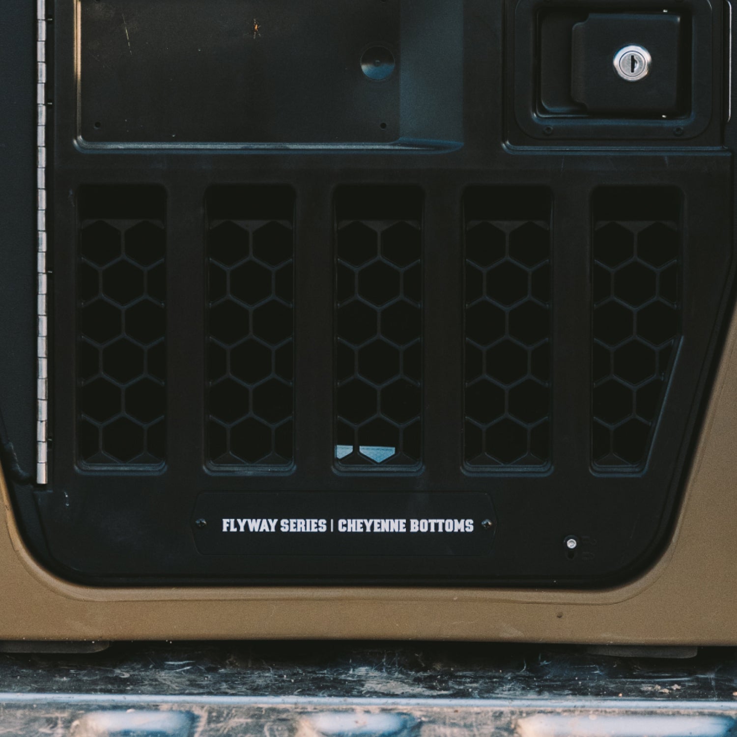 A tan and black Kennell dog crate sits in the back of a pickup truck, surrounded by hunting gear including a hanging deer head and other equipment. A door tag reads Flyway Series | Cheyenne Bottoms