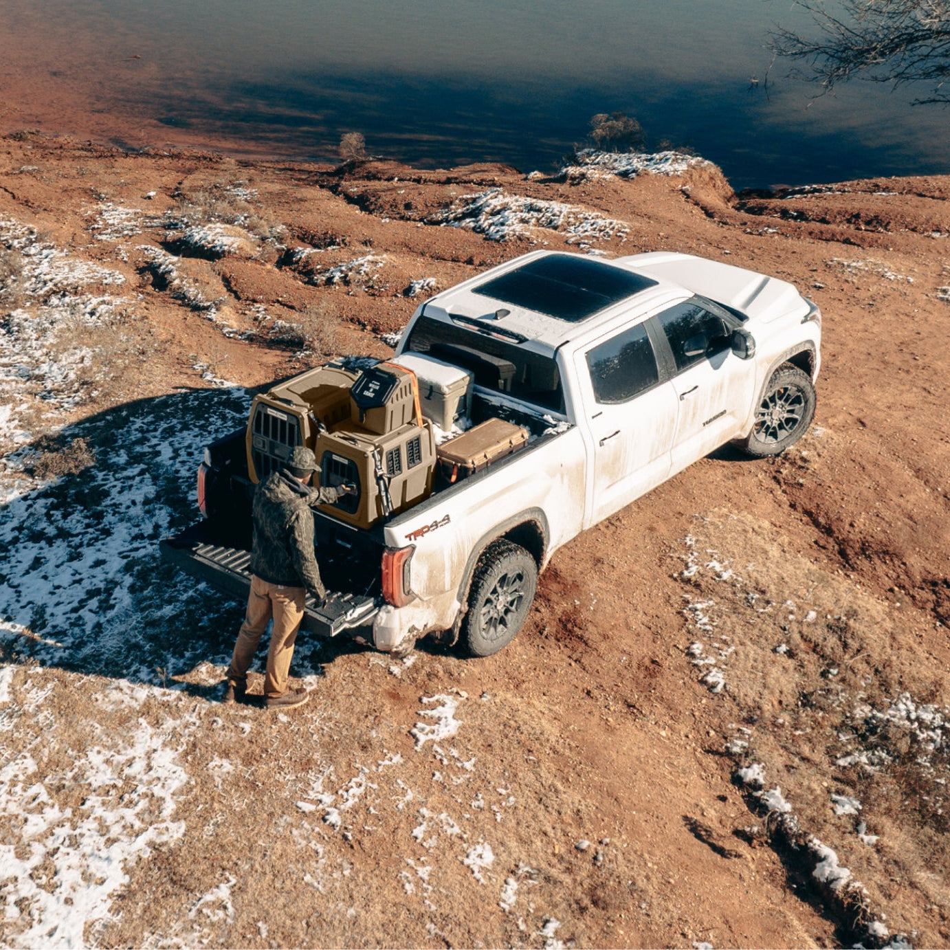 A white pickup truck parked on a snowy, rocky outdoor hillside, with a person standing in the open bed of the truck and loading or unloading gear. The scene is captured from above, with a dark body of water in the background.