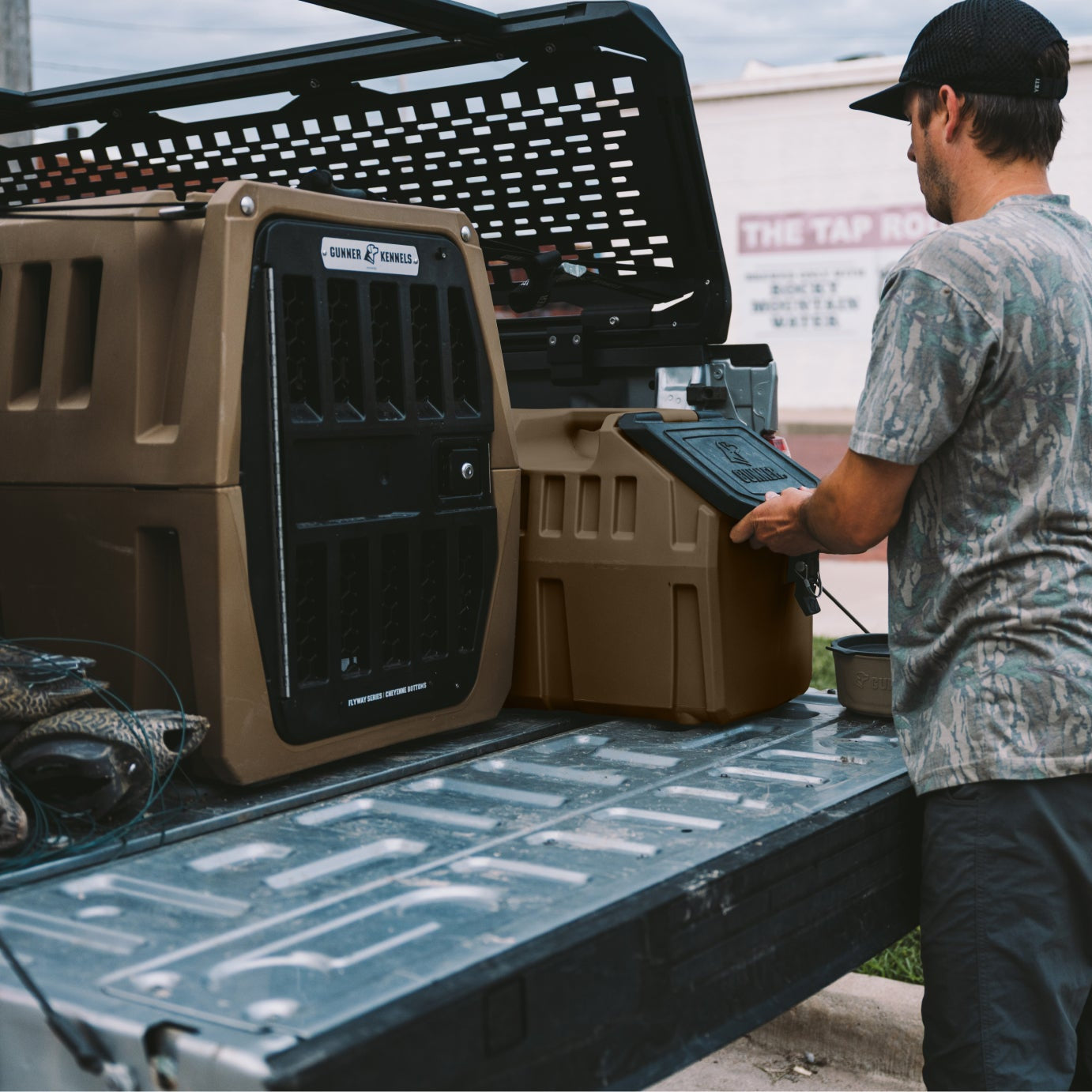 A man in a camouflage shirt and black cap stands in the bed of a truck next to a tan and black portable generator, holding a tablet. Another tan generator is parked beside him on a paved outdoor area.