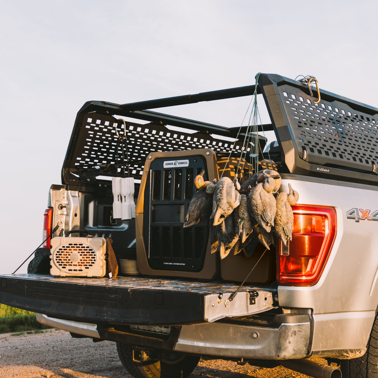 A black truck bed rack and cooler system installed in the tailgate of a silver pickup truck, with hunting ducks hanging from the side and a small speaker on the tailgate, photographed outdoors at sunset.