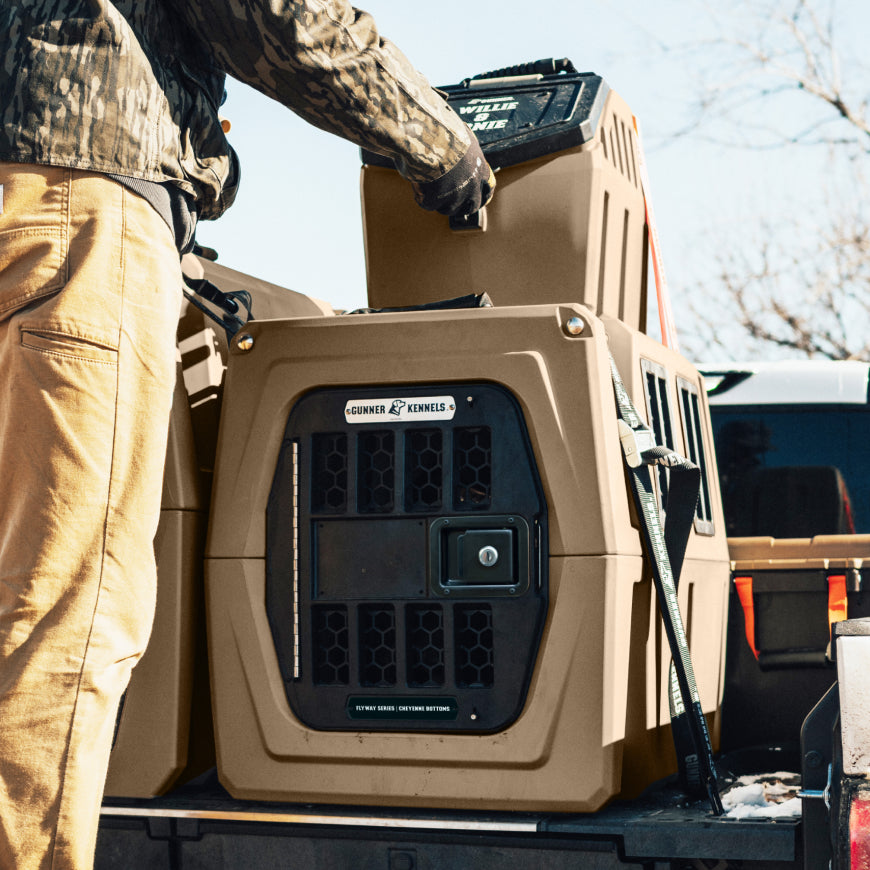 A tan and black dog crate secured in the bed of a pickup truck, with a person in camouflage clothing adjusting the top latch. The brand name 'CORNETS' is visible on the front of the crate.