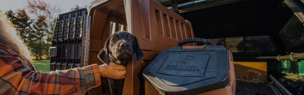 A person in a plaid shirt holds a black hunting dog inside a brown dog crate in the back of a vehicle, with a Gunner branded container nearby and trees visible in the background.