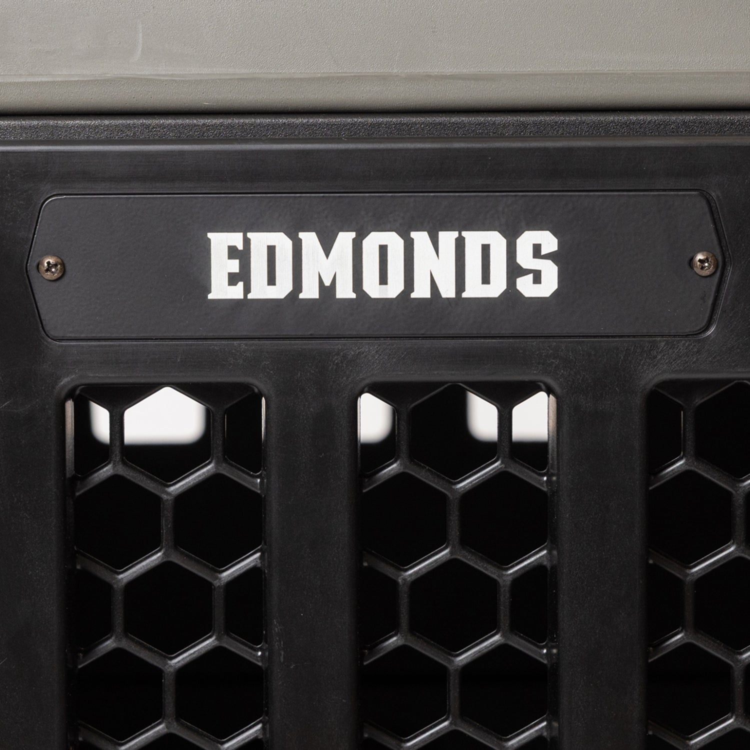 A black PERSONALIZED KENNEL DOOR TAG by GUNNER featuring a hexagonal grid and bold white EDMONDS lettering, resembling a custom dog tag, and attached with two screws.