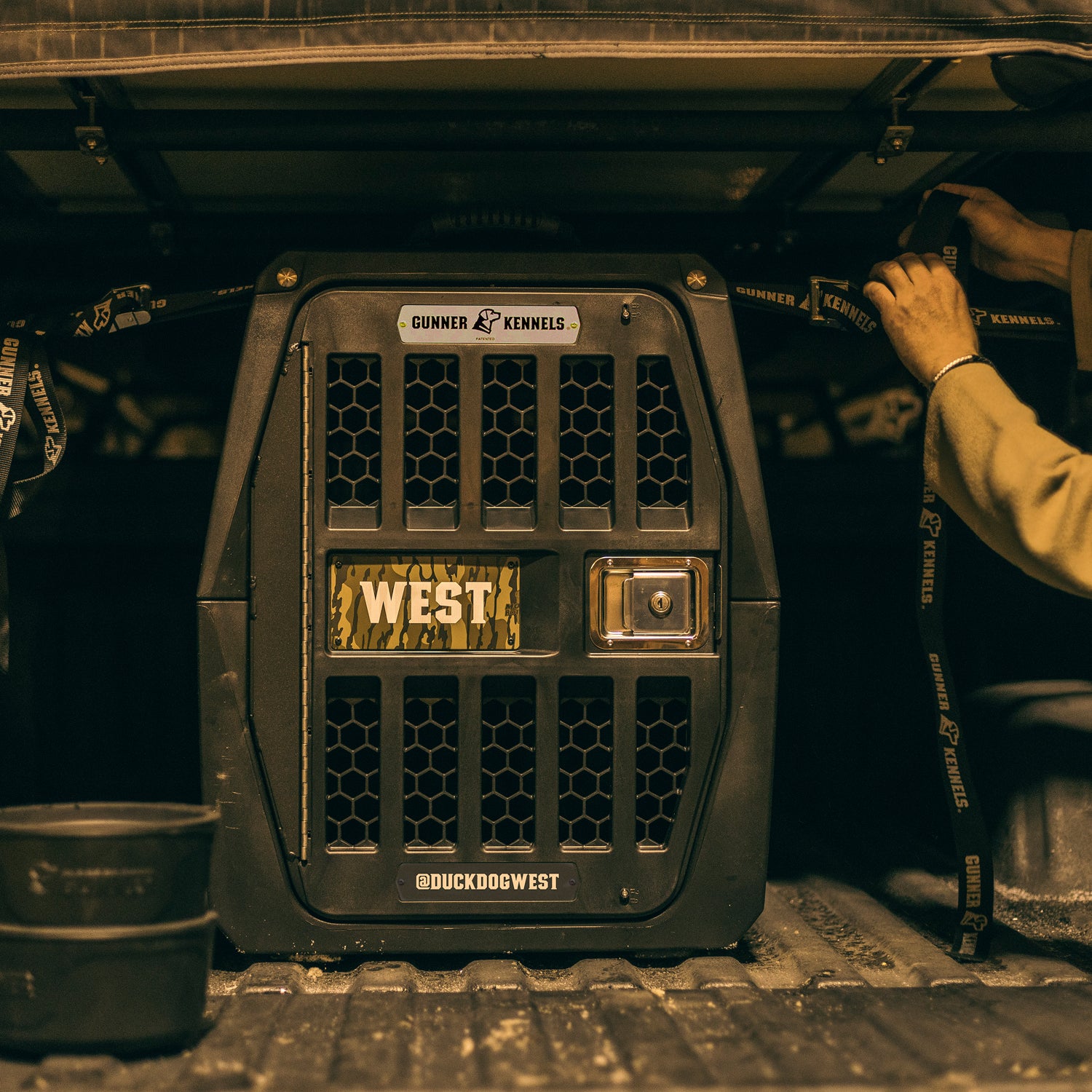 A sturdy black GUNNER crate sits in the back of a truck, with a person’s hands securing straps nearby. The crate features kennel branding and convenient carrying handles, while a PERSONALIZED KENNEL DOOR TAG is attached to the front, providing an extra touch of care for your pet’s identification.