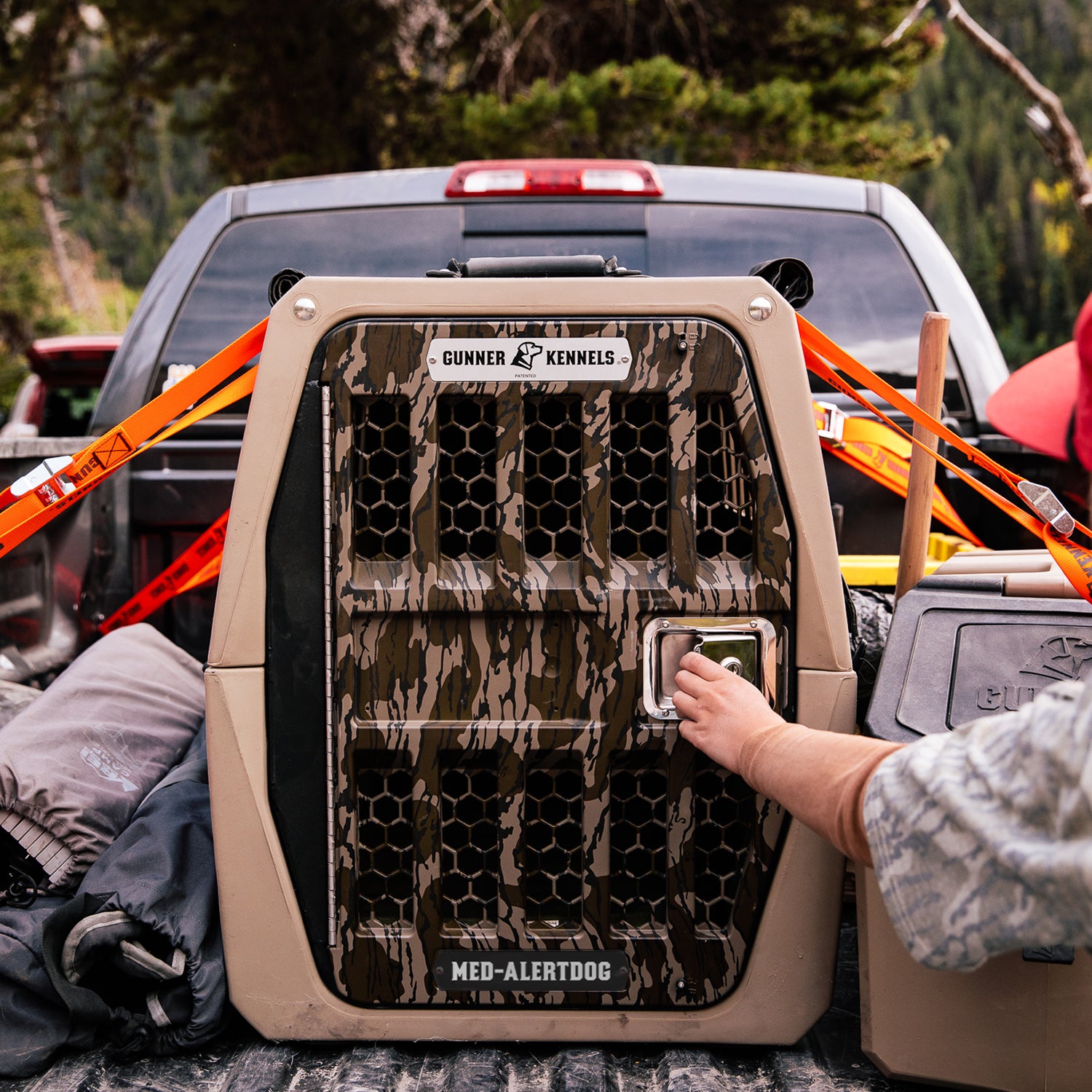 A person opens a camo-patterned dog crate labeled Gunner Kennels and Med-Alert Dog in the back of a pickup truck outdoors. Orange straps secure the crate, and a personalized dog tag hangs from the dog’s collar, ready for any adventure.