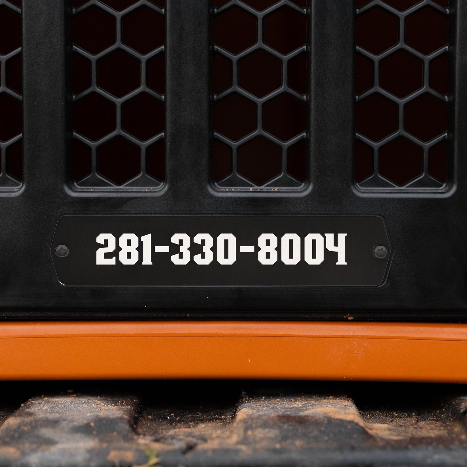 Black nameplate on orange heavy equipment grille resembles an engraved dog tag, displaying phone number 281-330-8004 in white bold text, with hexagonal mesh grille pattern above and rubber track below.