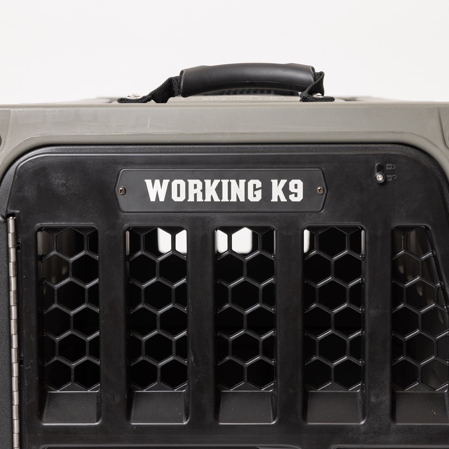 A close-up of a black and gray dog crate with a handle on top and a WORKING K9 label above hexagonal ventilation holes, featuring a GUNNER PERSONALIZED KENNEL DOOR TAG attached for easy identification.