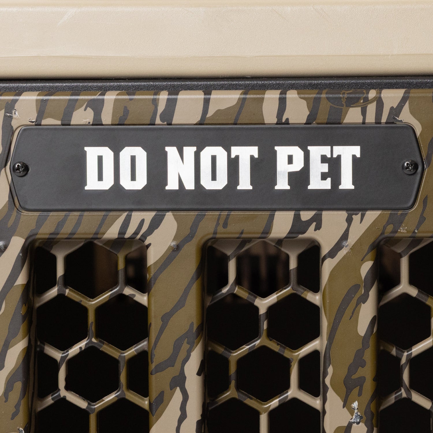 A camouflage-patterned crate from GUNNER featuring a striking black sign with bold white “DO NOT PET” lettering above a honeycomb-patterned vent, complemented by a PERSONALIZED KENNEL DOOR TAG for added customization.