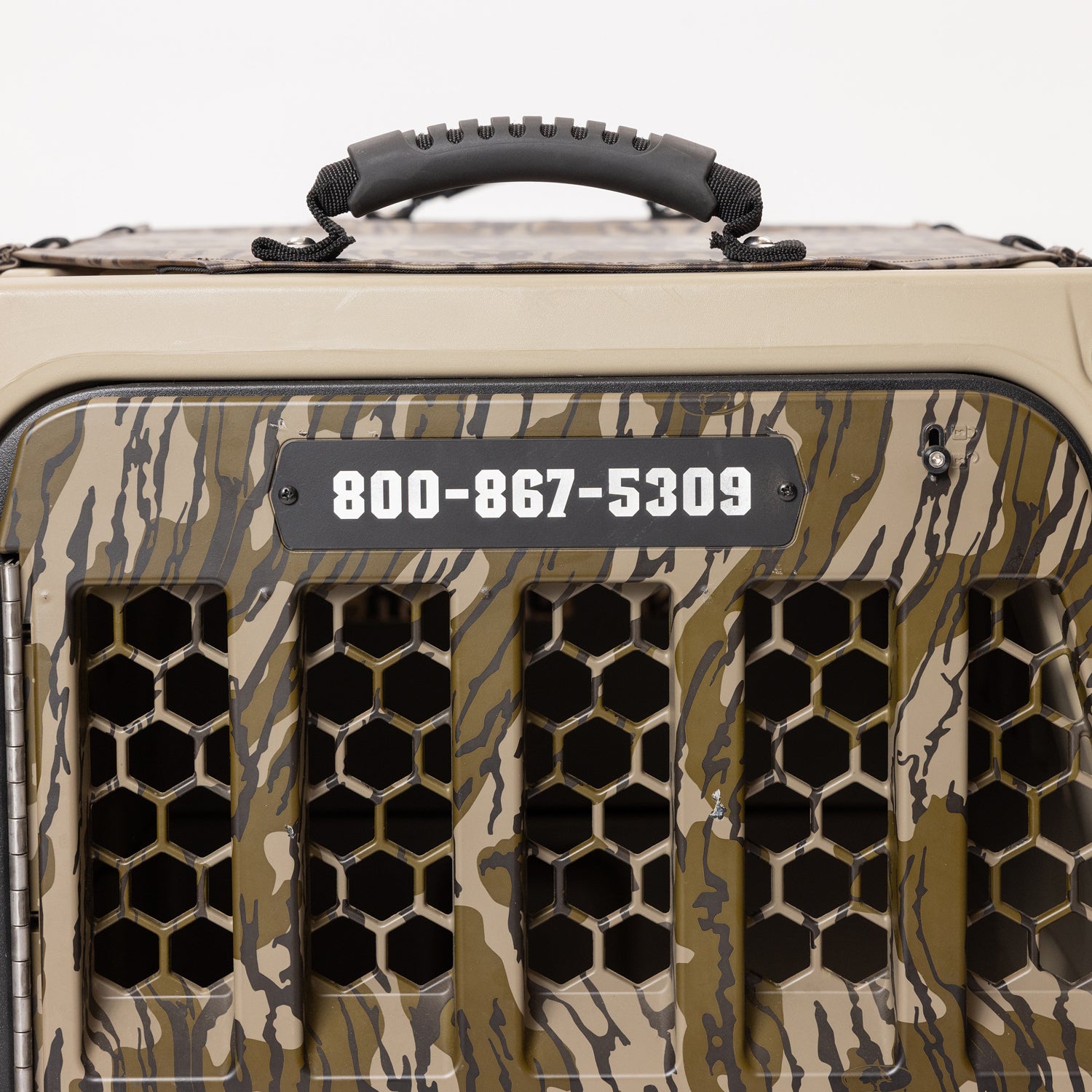 Close-up of a GUNNER kennel featuring a camouflage pattern, black handle, and honeycomb mesh door. A black PERSONALIZED KENNEL DOOR TAG is visible, displaying the phone number 800-867-5309—providing crucial identification for your pet, just like an engraved dog tag.