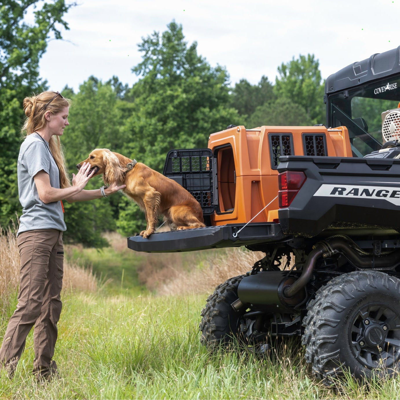 Person interacting with a dog and a GUNNER kennel on the back of a Ford Ranger vehicle in a natural setting.