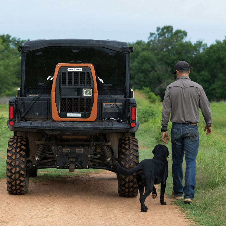 A man in a gray shirt and cap walks his black dog beside an off-road vehicle carrying the GUNNER G1™ KENNEL - Ducks Unlimited Orange on the back, showing his Ducks Unlimited spirit along a dirt road surrounded by greenery.
