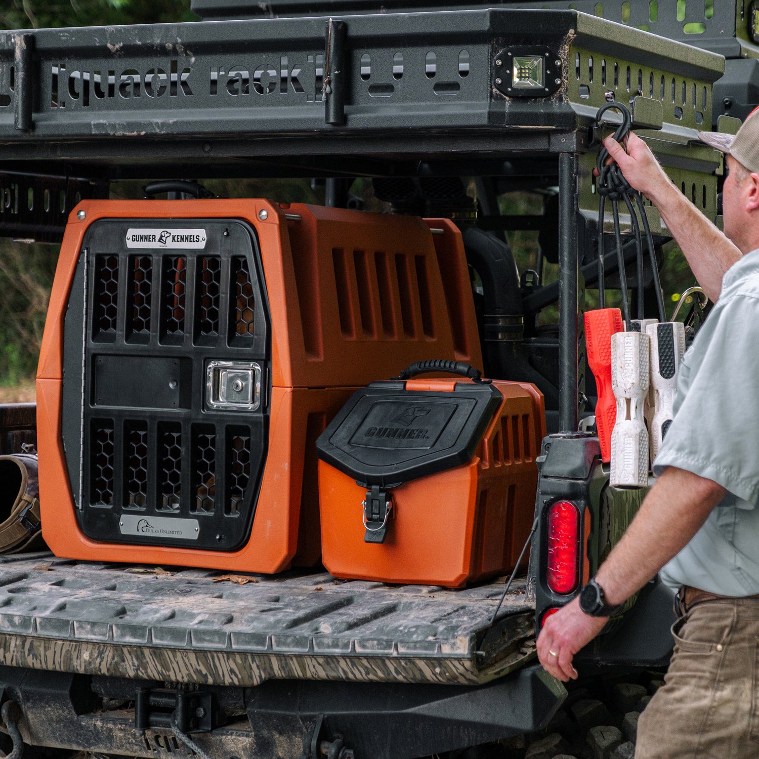 A person loads a GUNNER FOOD CRATE - Ducks Unlimited Orange and matching waterproof gear onto an off-road utility vehicle, with various GUNNER accessories like an airtight dog food storage container and training dummies visible.