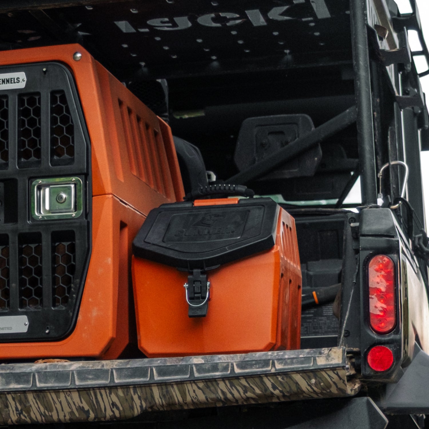 A GUNNER FOOD CRATE - Ducks Unlimited Orange is strapped in the back of an off-road vehicle, its airtight black lid visible. Next to it sits a larger GUNNER kennel, ideal as a waterproof dog food storage container. Mud splashes the rugged bumper.