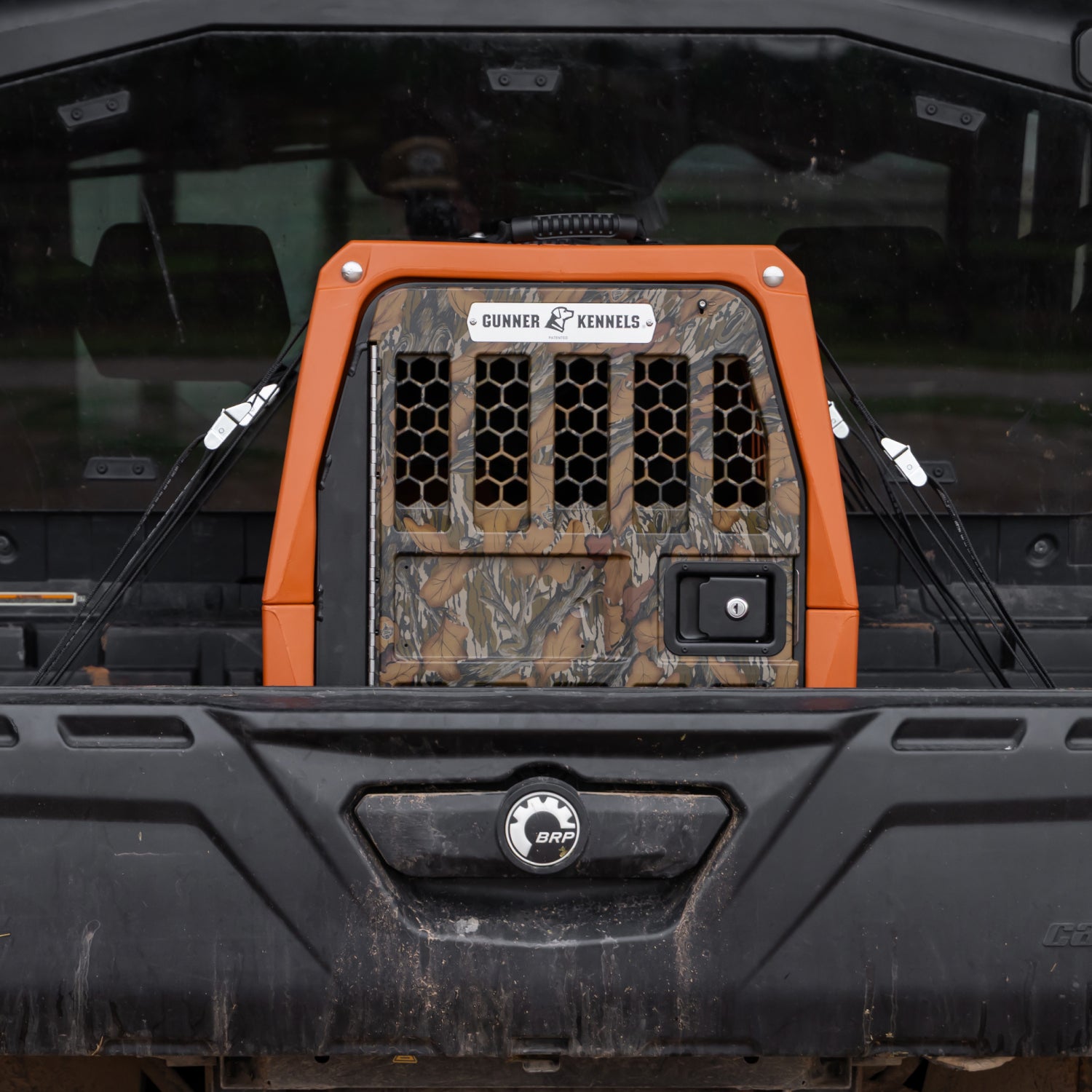 A GUNNER G1™ KENNEL - Habitat Edition with a camouflage pattern and small ventilation holes is secured in the bed of a black BRP utility vehicle, view from the rear. The crate features a label: Gunner Kennels x Ducks Unlimited.