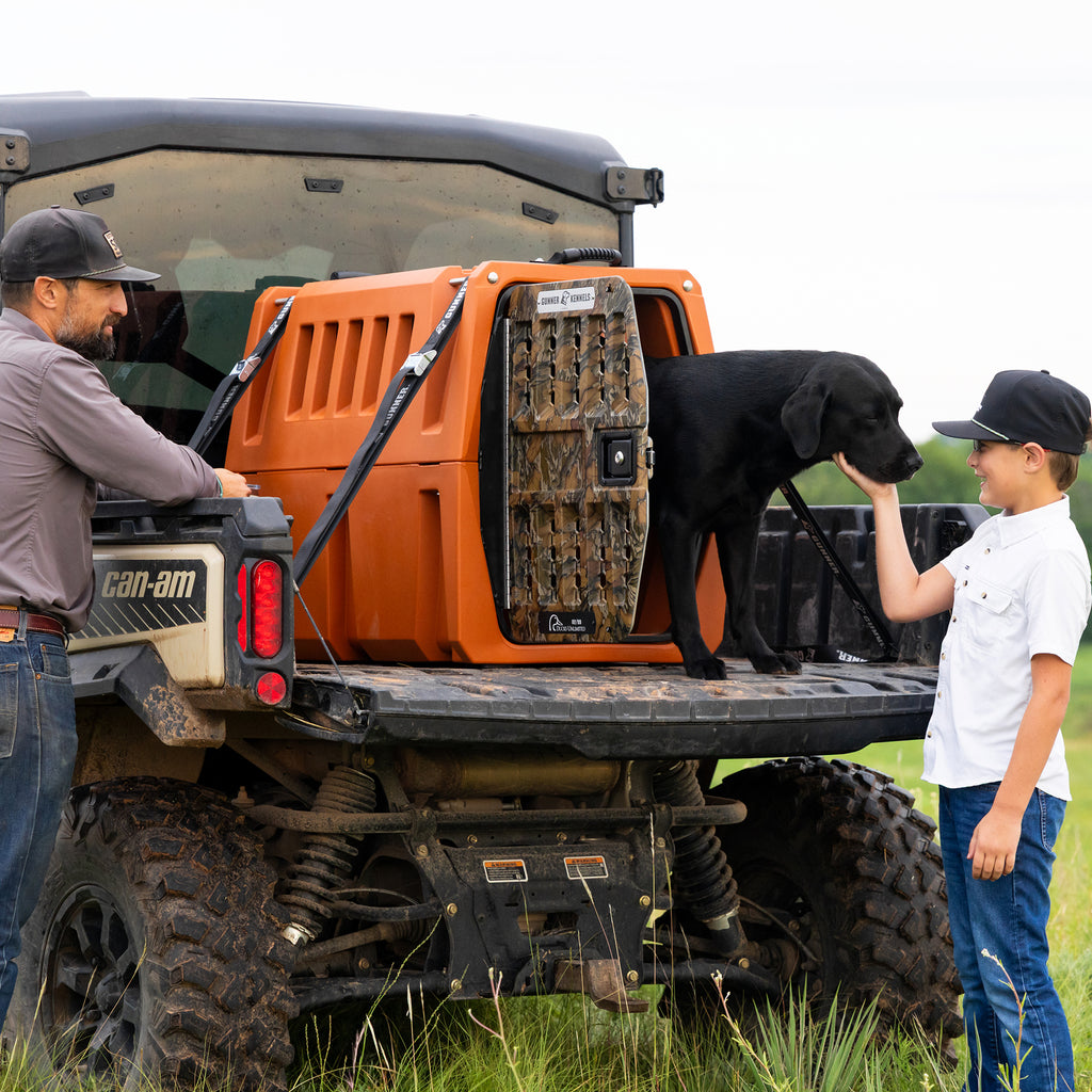 A man and boy stand by an off-road vehicle with a GUNNER G1™ KENNEL - Habitat Edition in the back. A black dog at the edge sniffs the boy’s hand as they smile at each other in a grassy outdoor area.