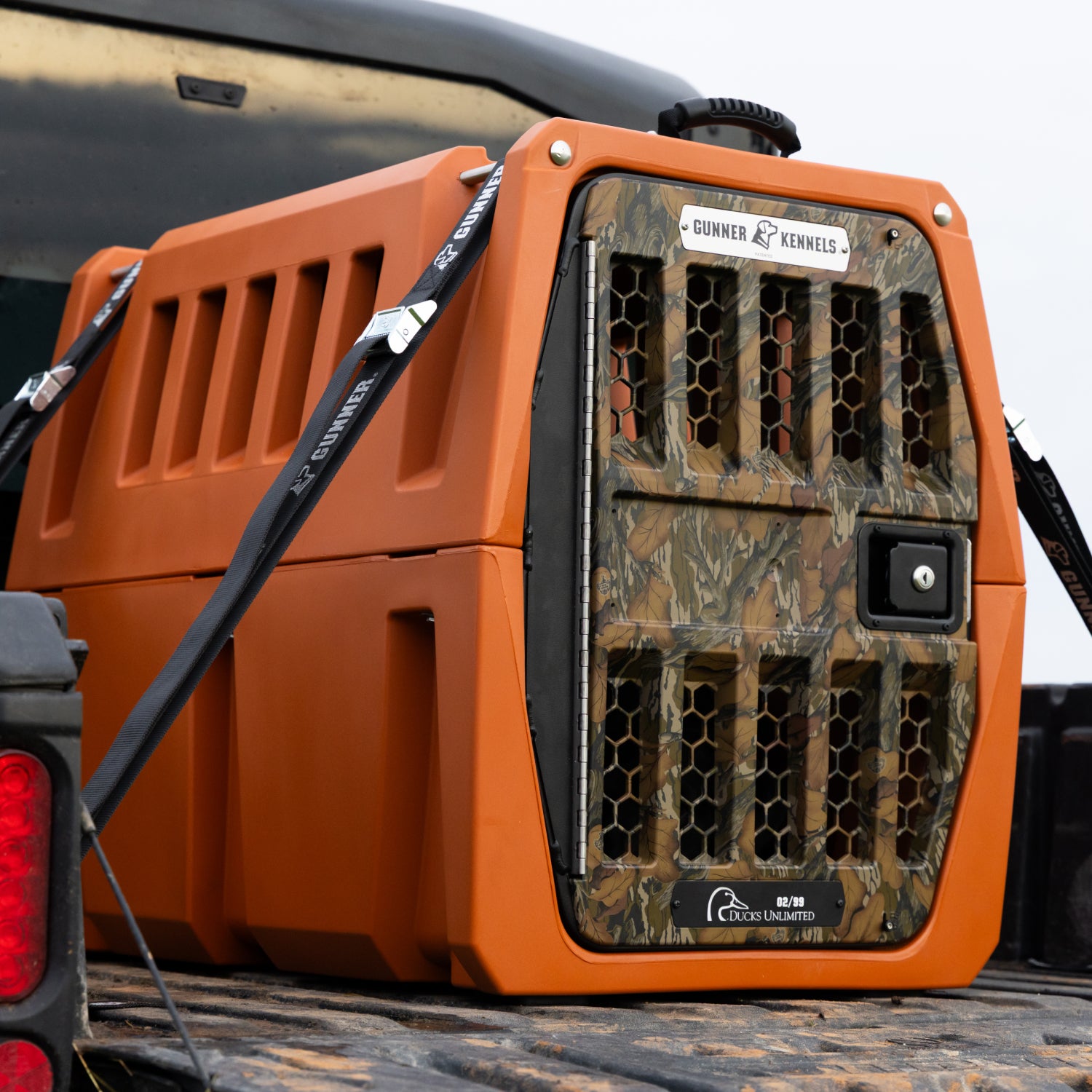 A GUNNER G1™ KENNEL - Habitat Edition, featuring an orange double-wall build, camouflage door, ventilation slats, black straps, and a Ducks Unlimited decal, is secured in a pickup truck bed.