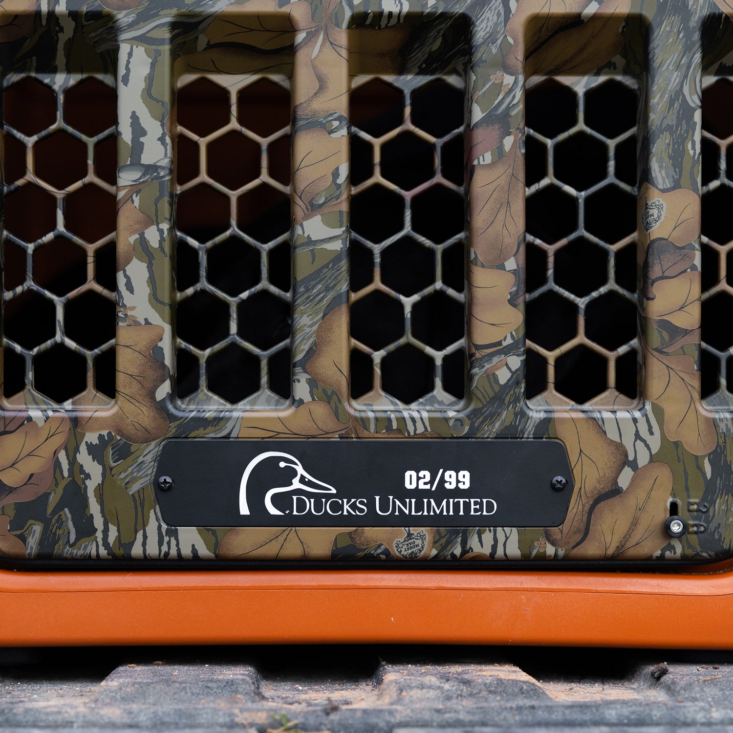 A close-up of the GUNNER G1™ KENNEL - Habitat Edition, featuring a camouflage pattern, hexagonal vents, orange base, and DUCKS UNLIMITED 02/99 metal plate with a duck logo—engineered as the safest dog crate for your companion.
