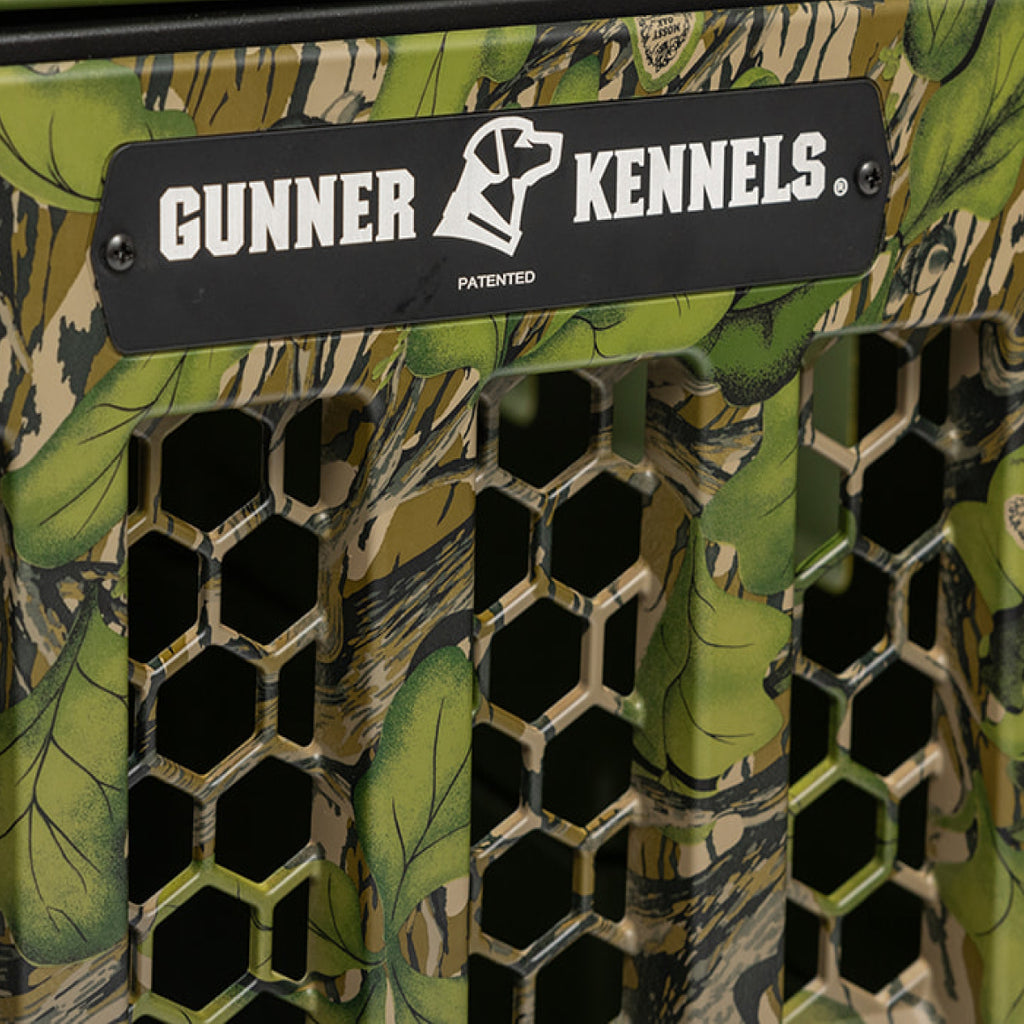 Close-up of the GUNNER G1™ Kennel Door in Mossy Oak Full Foliage, showing hexagonal ventilation holes and a black nameplate with the dog logo and Gunner branding.