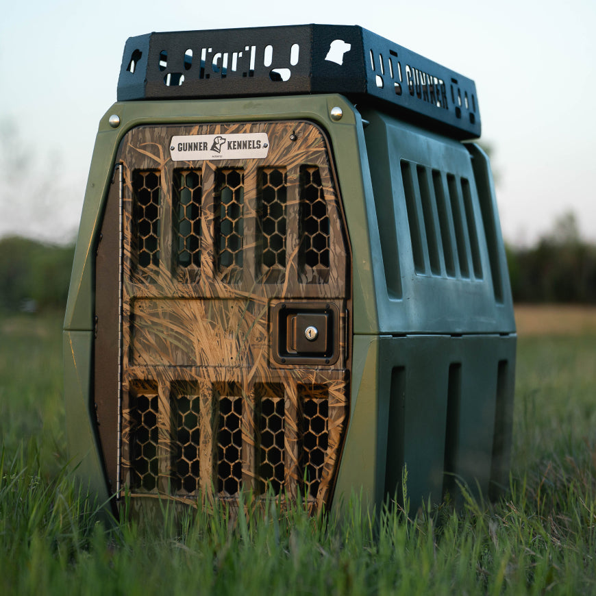 The GUNNER G1™ KENNEL in green/mossy oak shadow grass sits on grass outdoors, featuring a honeycomb metal door and reinforced corners. This double-walled crate is the safest travel kennel for pets, proudly made in America.