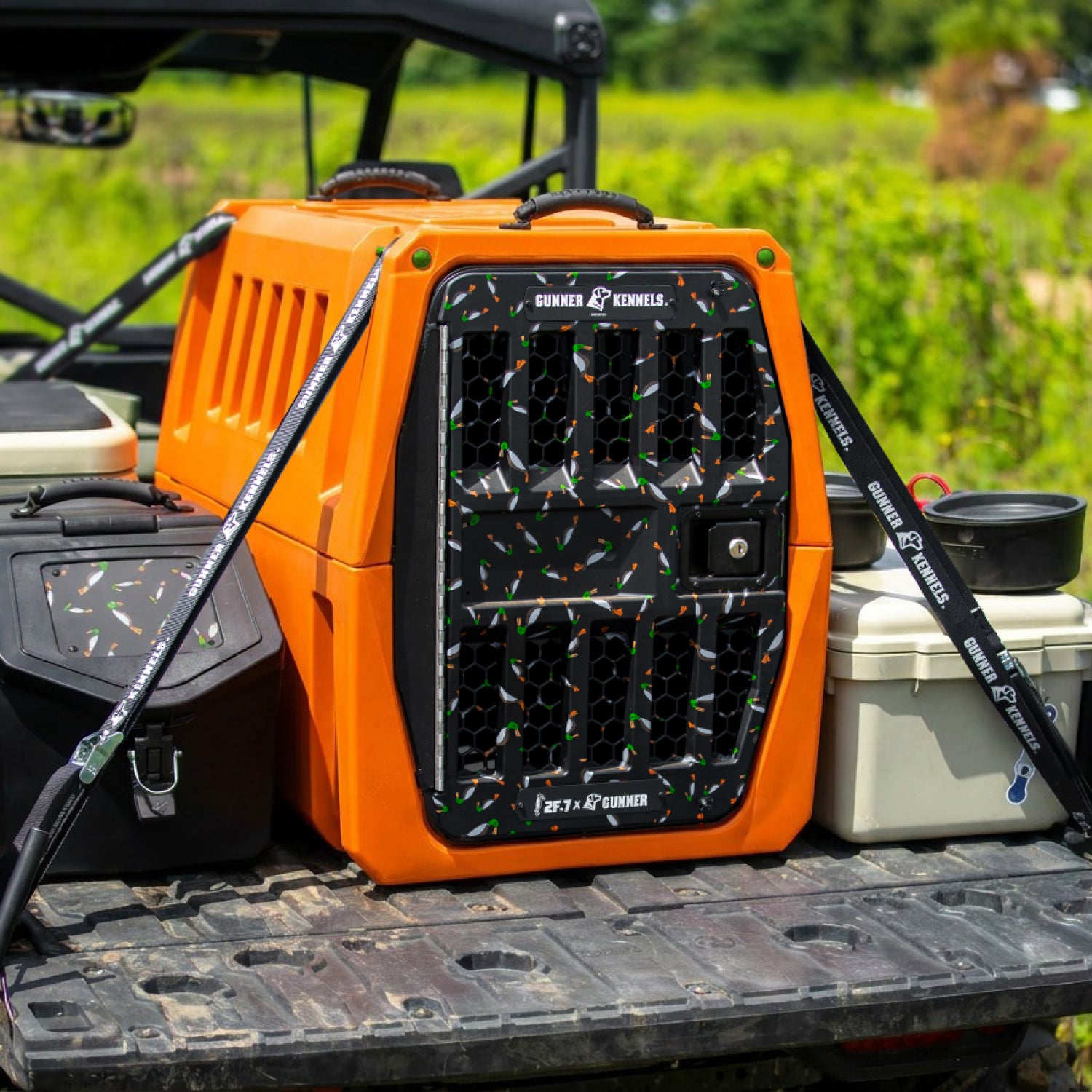 The GUNNER G1™ KENNEL - 2F.7 ORANGE CRUSH, featuring a double-walled design and shock protection, sits in a pickup bed with its black speckled door facing out, surrounded by gear and storage boxes against a green backdrop.