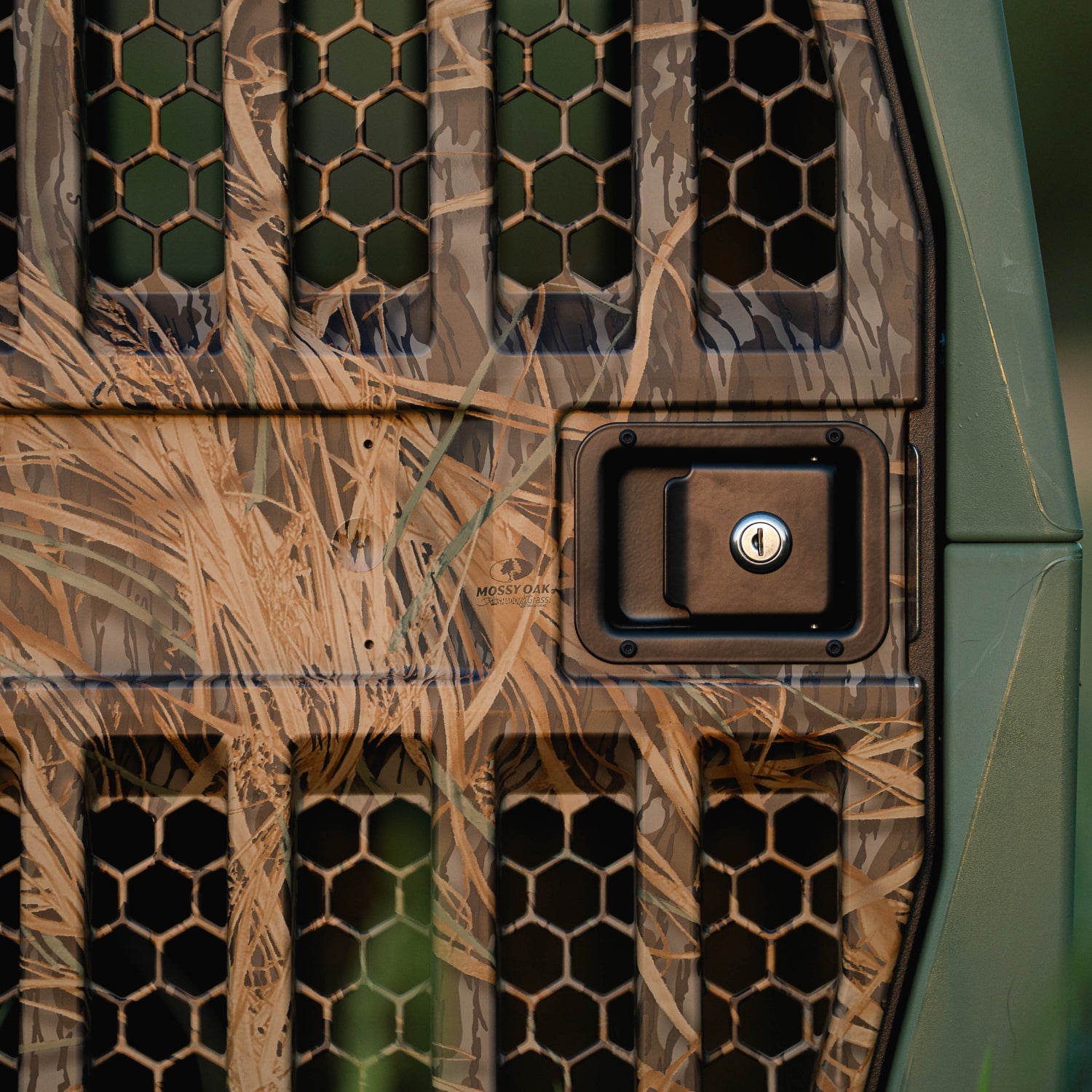 Close-up of a vehicle door with grass and leaf camouflage, hexagonal vents, and a rectangular handle—echoing the rugged design of the GUNNER G1™ KENNEL in Gunner Green/Mossy Oak Shadow Grass. Green and brown tones complete the look.